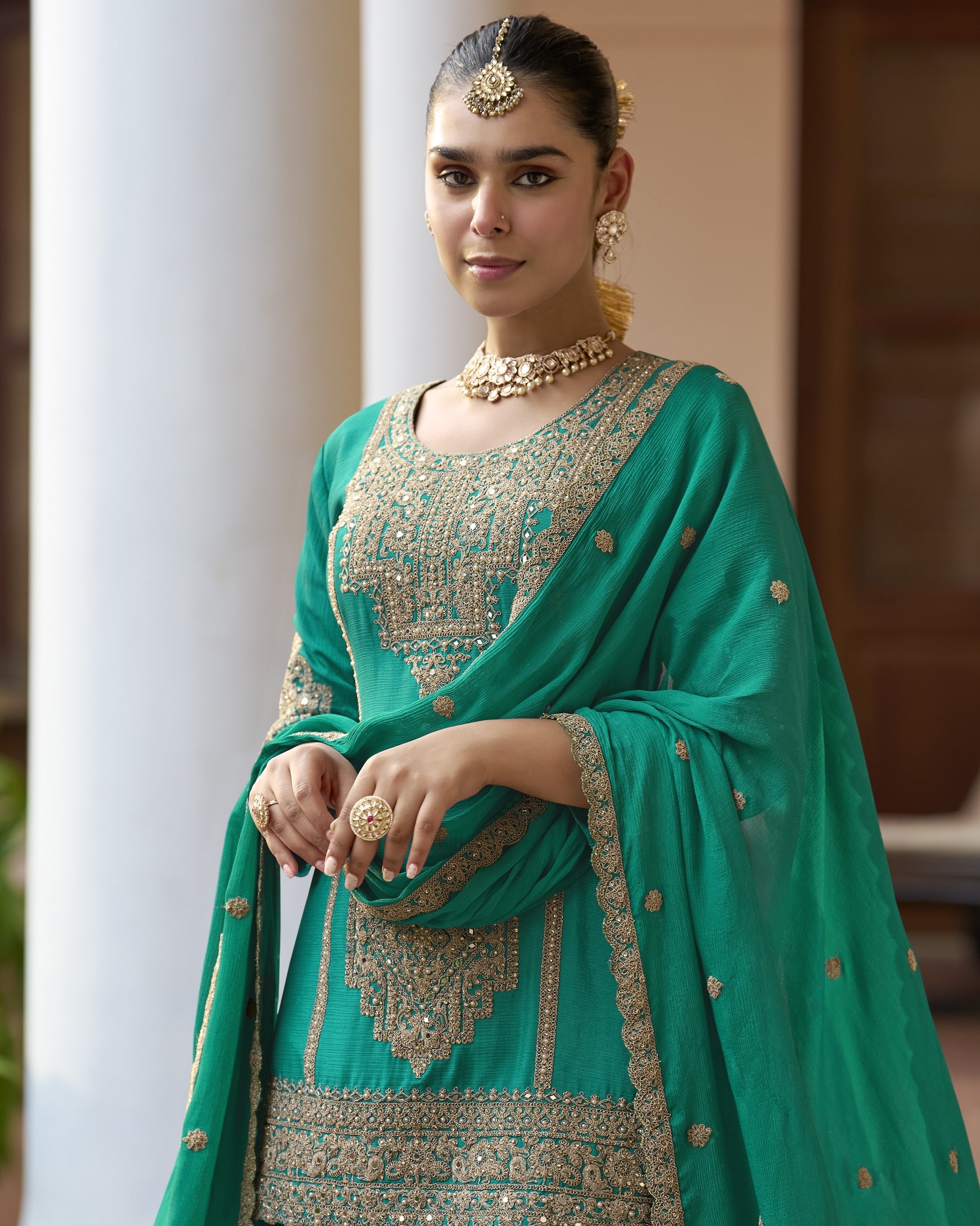 Woman in a teal traditional outfit with gold embroidery standing outdoors.