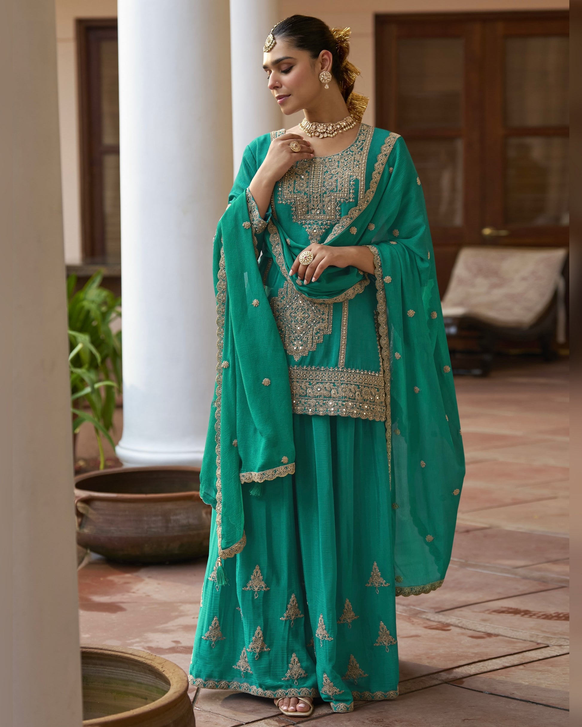 Woman in a teal traditional outfit with gold embroidery standing outdoors.
