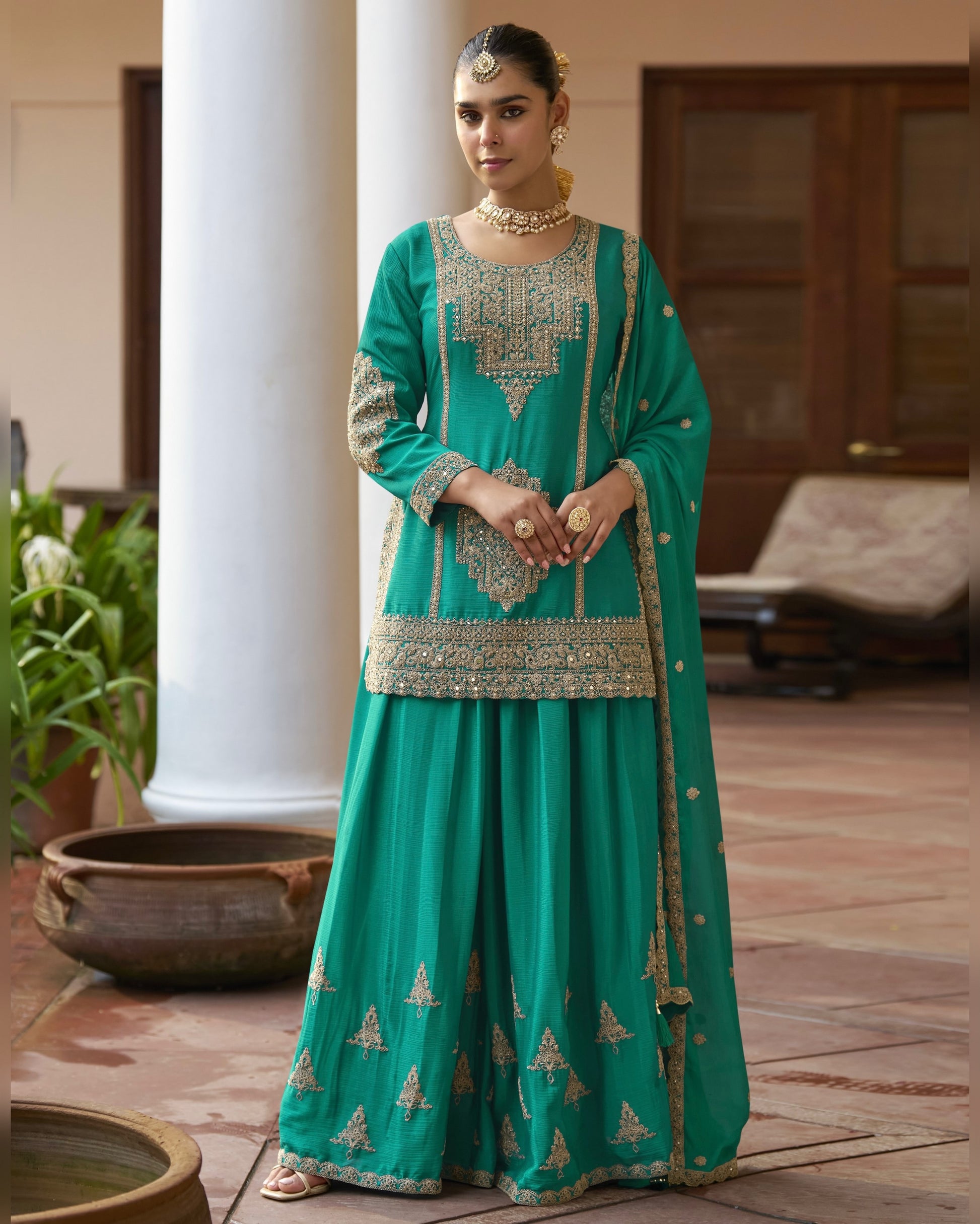 Woman in a teal traditional outfit with gold embroidery standing outdoors.