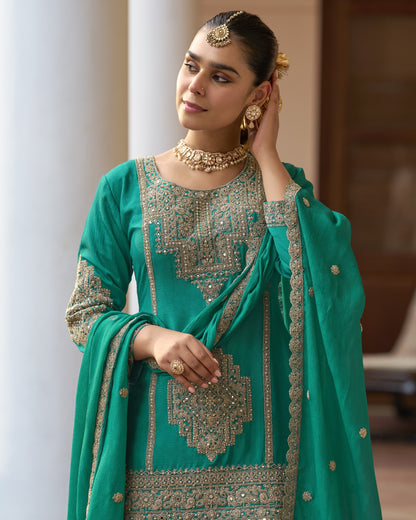 Woman in a teal traditional outfit with gold embroidery standing outdoors.