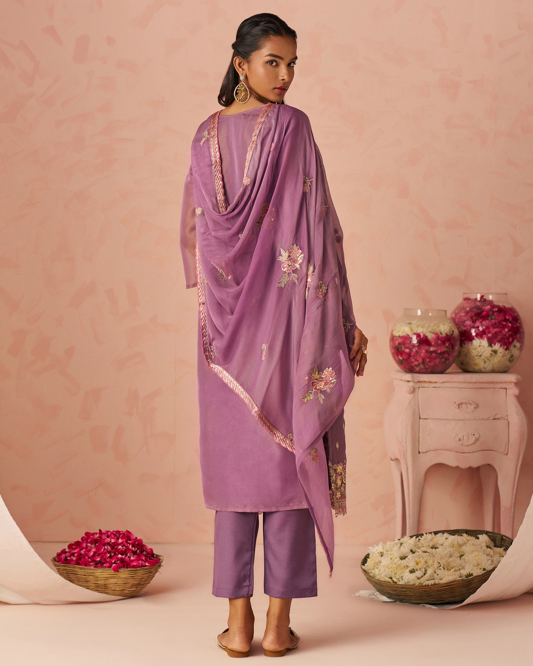 Woman wearing a purple traditional outfit with floral embroidery against a pink background.