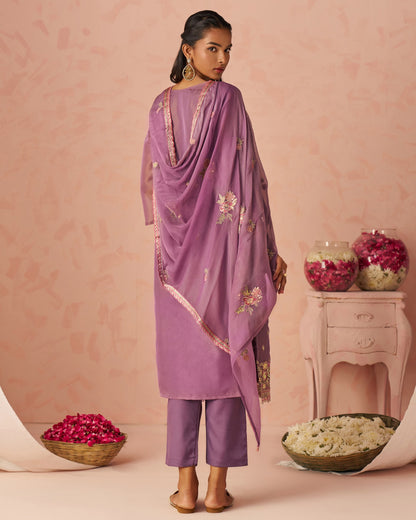 Woman wearing a purple traditional outfit with floral embroidery against a pink background.