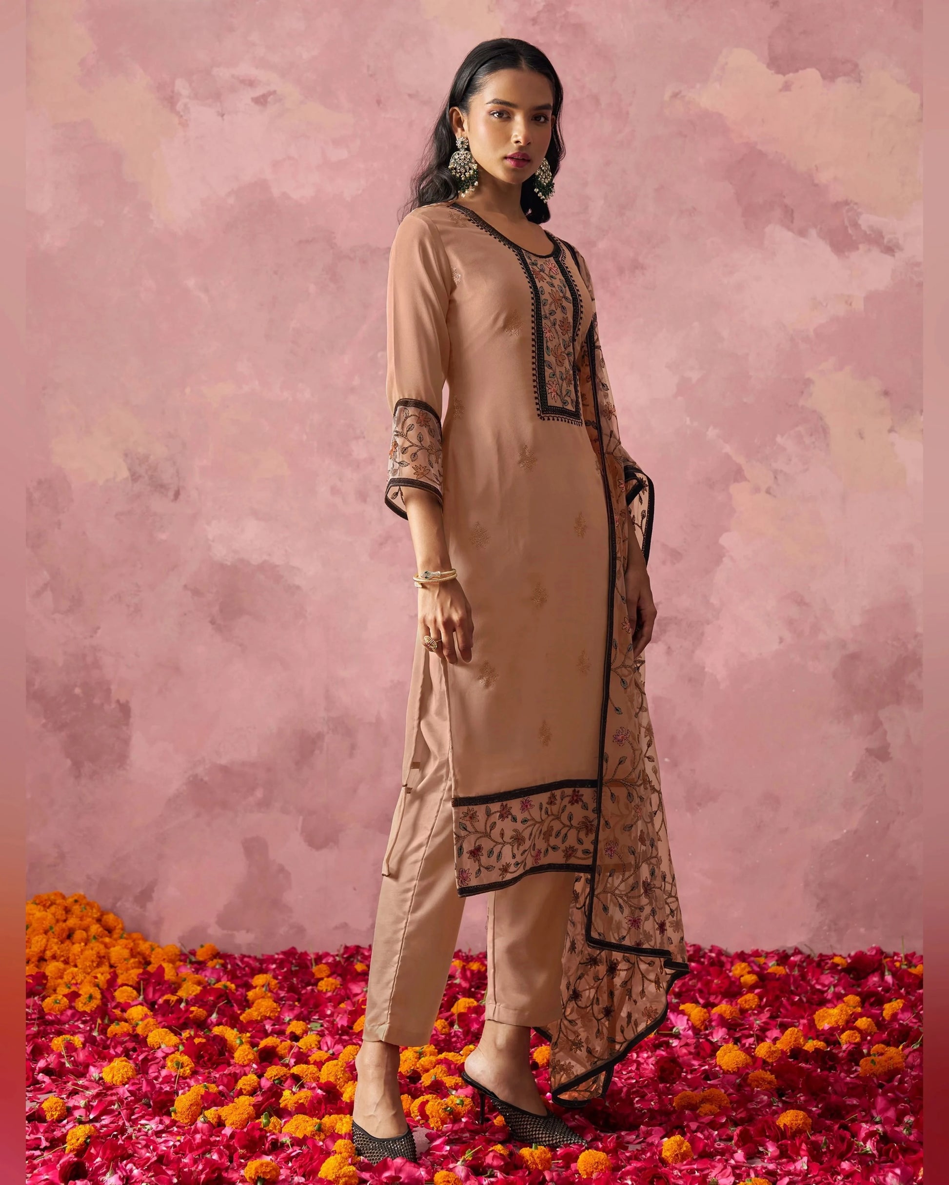 Woman in a beige and brown traditional outfit standing on flower petals with a pink floral background