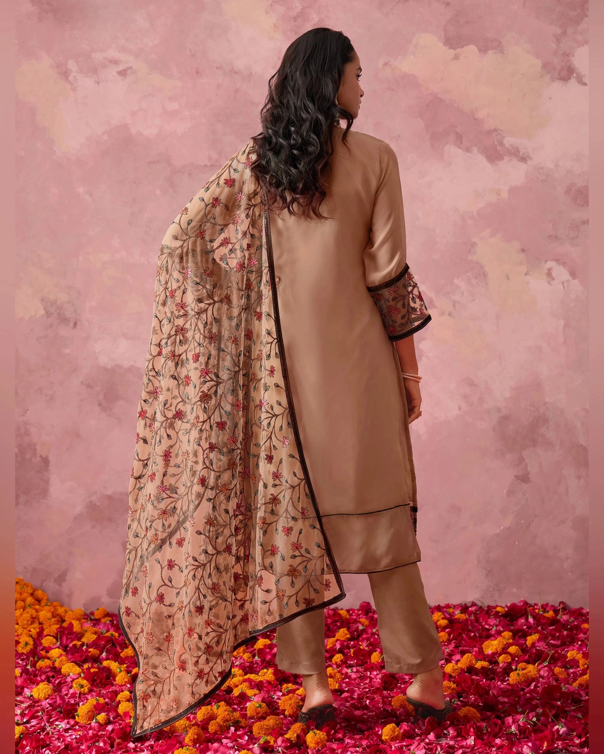 Woman in a beige traditional outfit with a floral dupatta standing on rose petals against a pink floral background