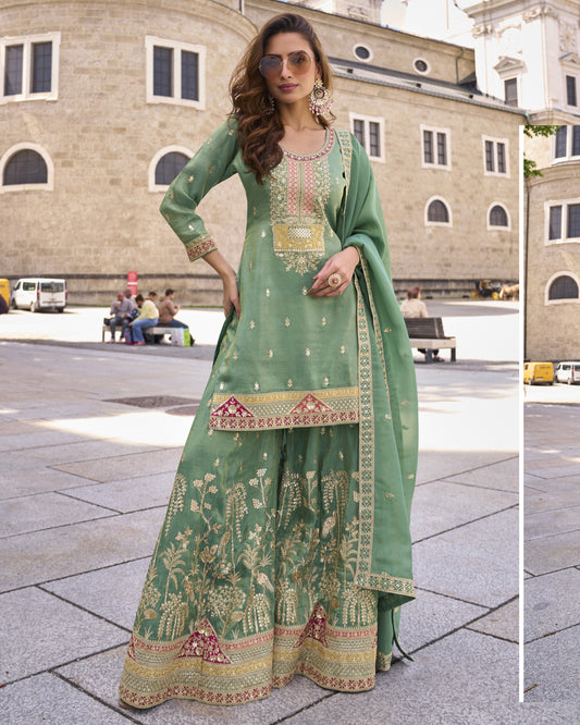 Woman in a green traditional outfit with intricate patterns standing in front of a building.