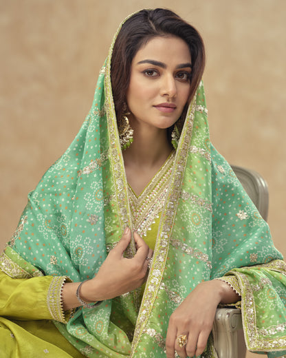 Woman wearing a green and yellow traditional outfit with a matching dupatta against a beige background