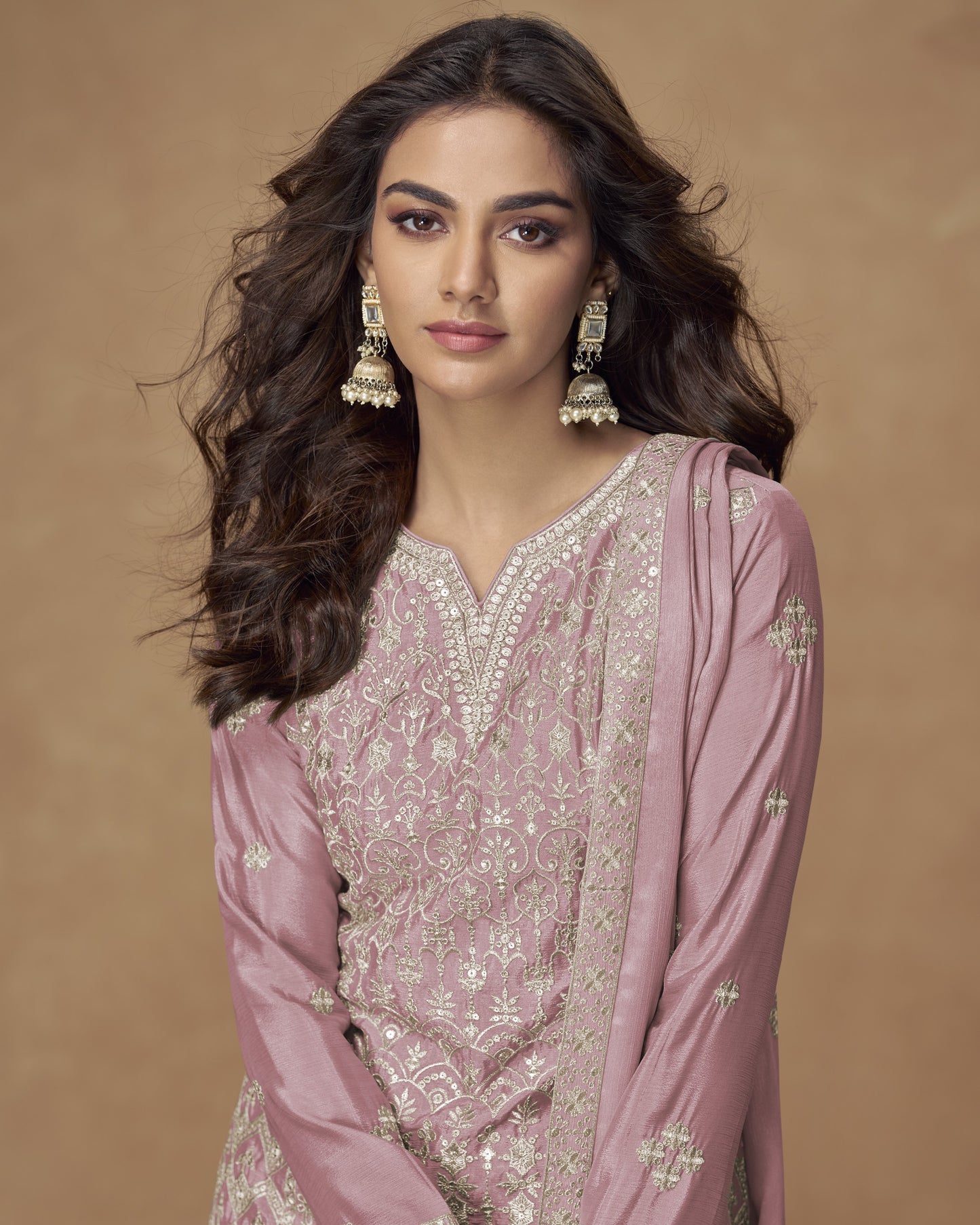 Woman wearing a pink embroidered traditional outfit with gold jewelry against a brown background