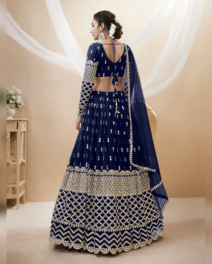 Woman wearing a blue and silver embroidered traditional outfit in a beige room.