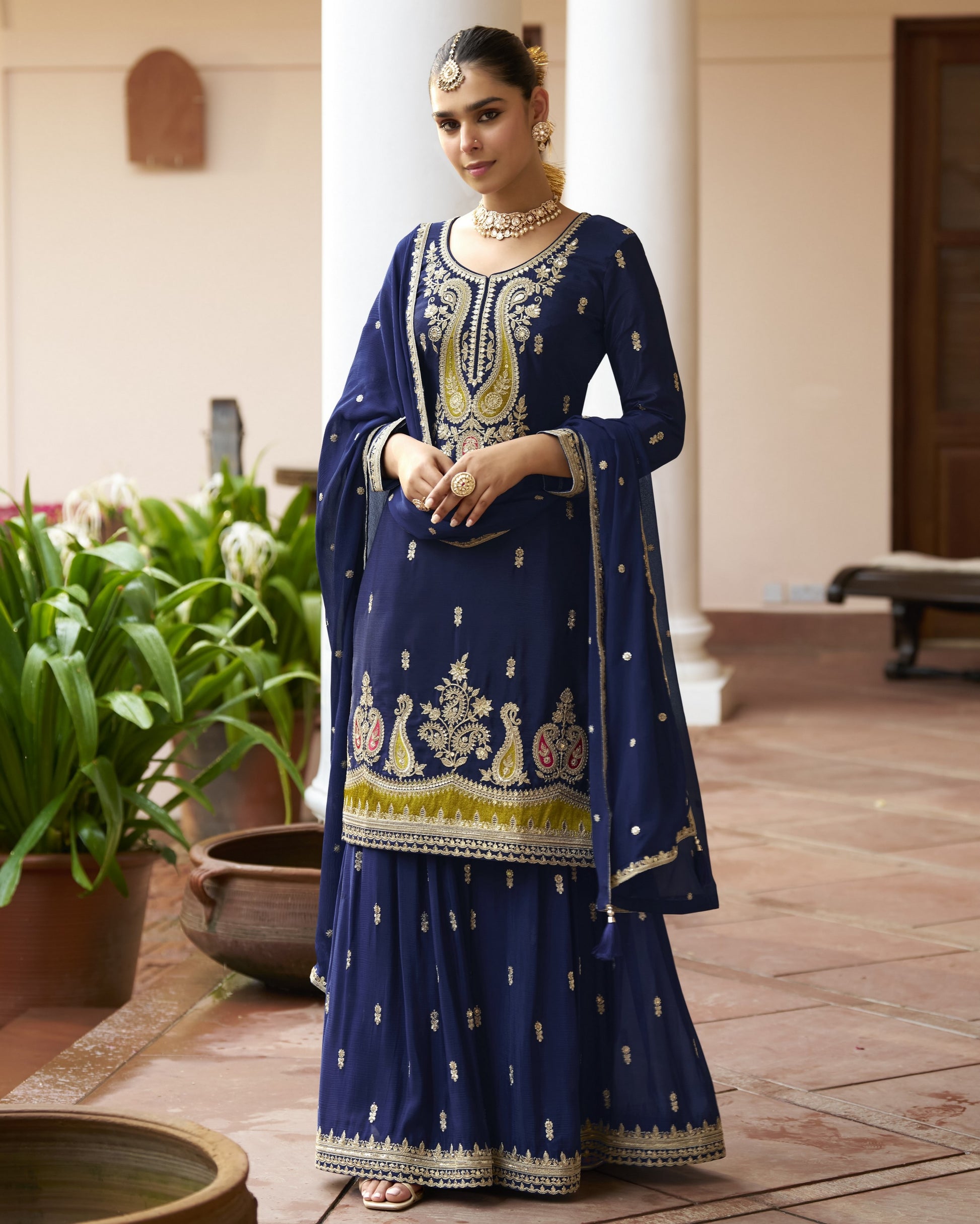 Woman in a traditional blue embroidered outfit standing in an outdoor setting with plants.
