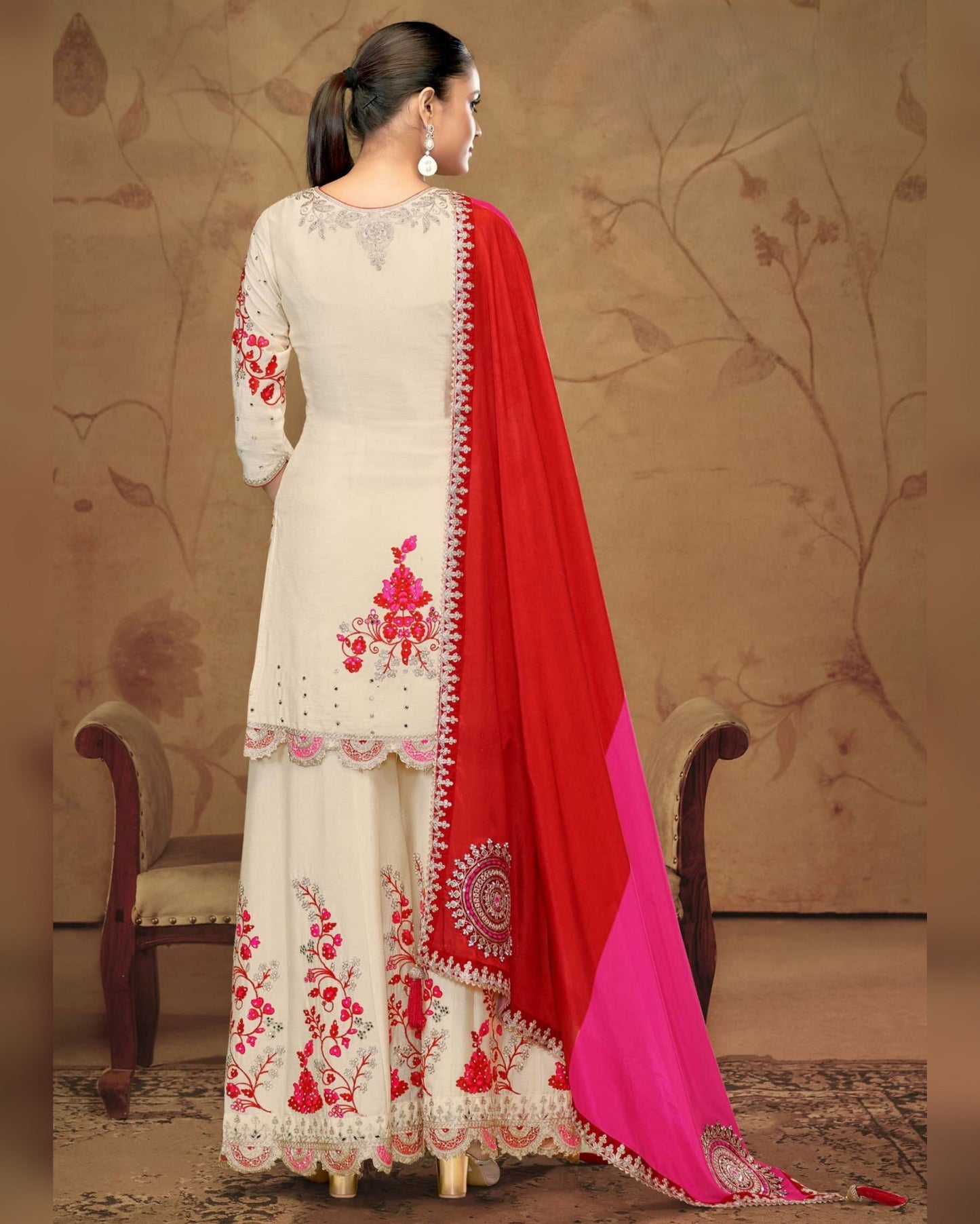 Woman wearing a white embroidered dress with red and pink dupatta against a beige wall.