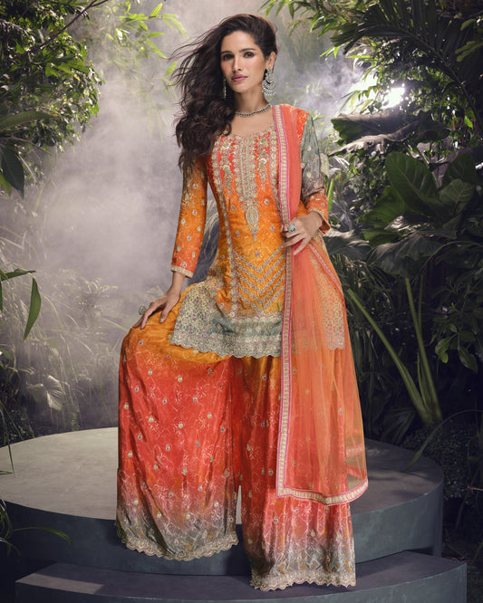 Woman in an orange and green traditional outfit with a floral pattern, sitting on a platform with greenery in the background.
