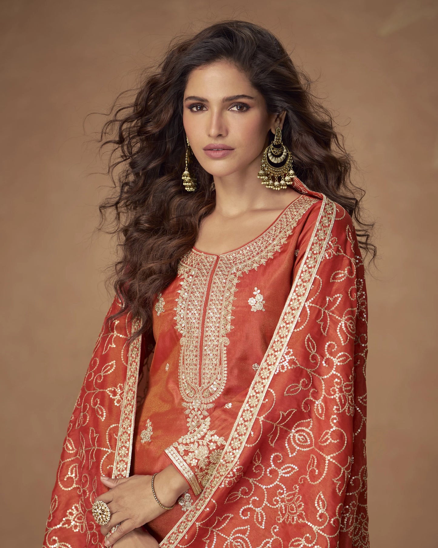 Woman wearing a traditional red embroidered outfit with gold jewelry against a brown background