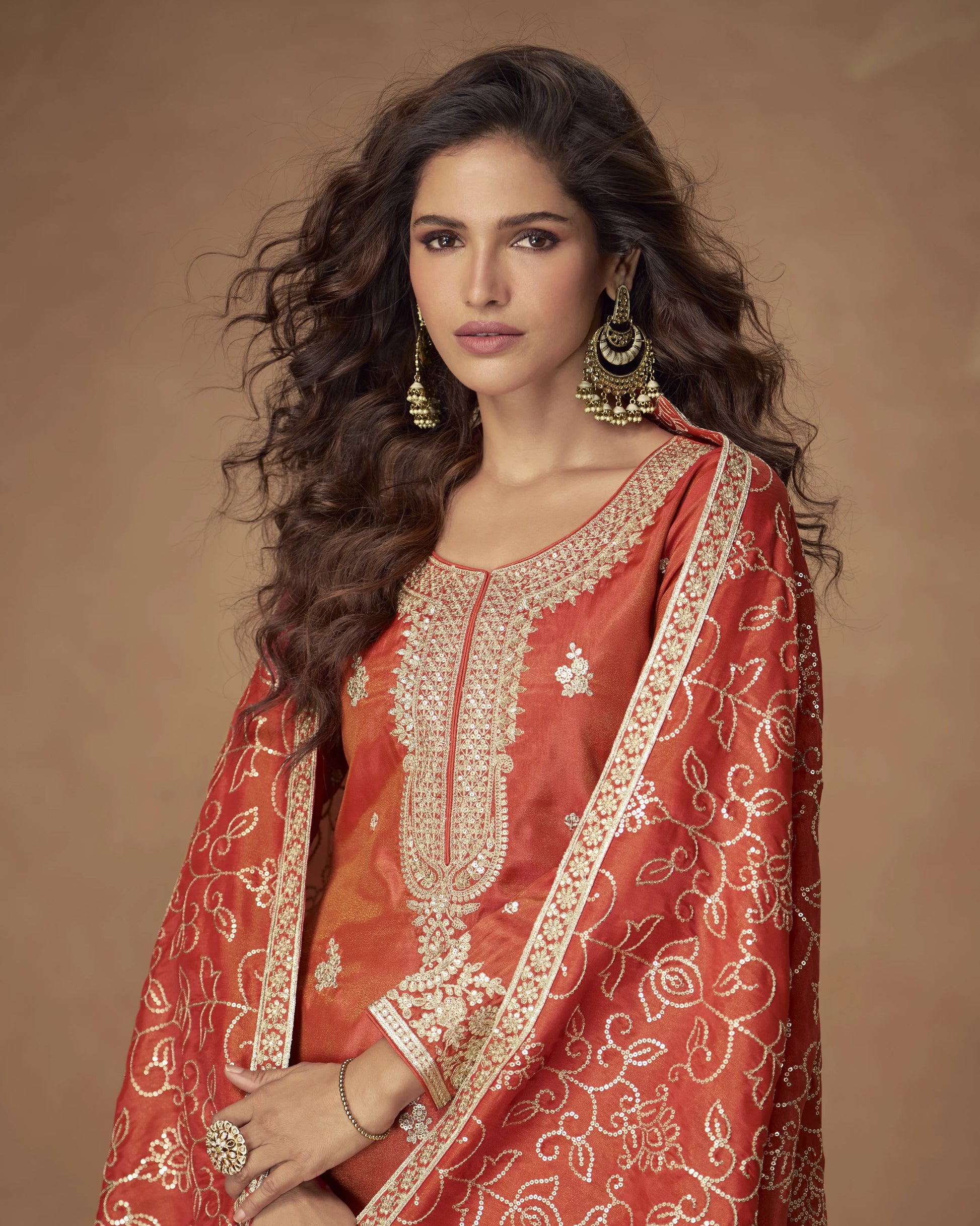 Woman wearing a traditional red embroidered outfit with gold jewelry against a brown background