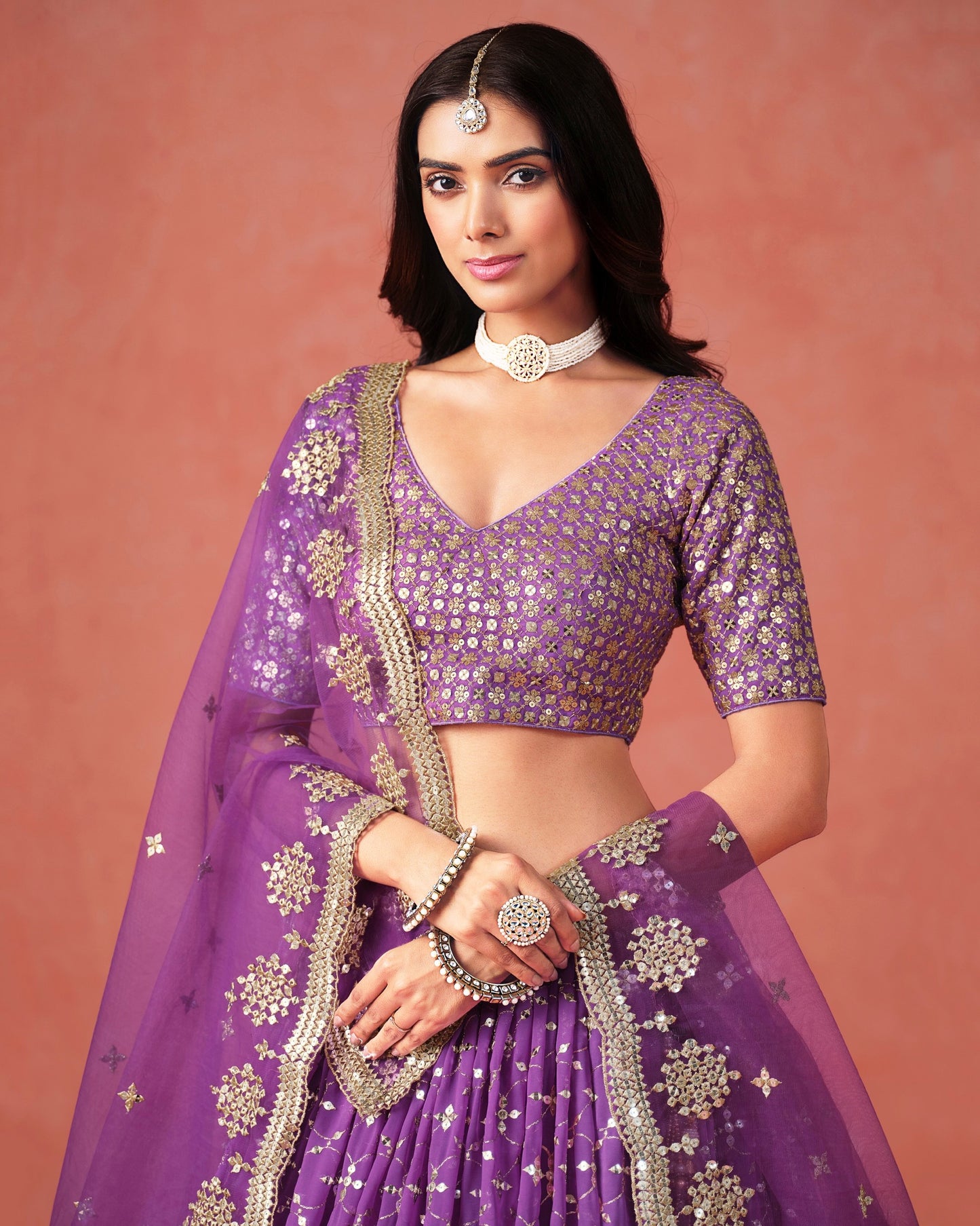 Woman wearing a purple traditional outfit with gold embroidery on a pink background