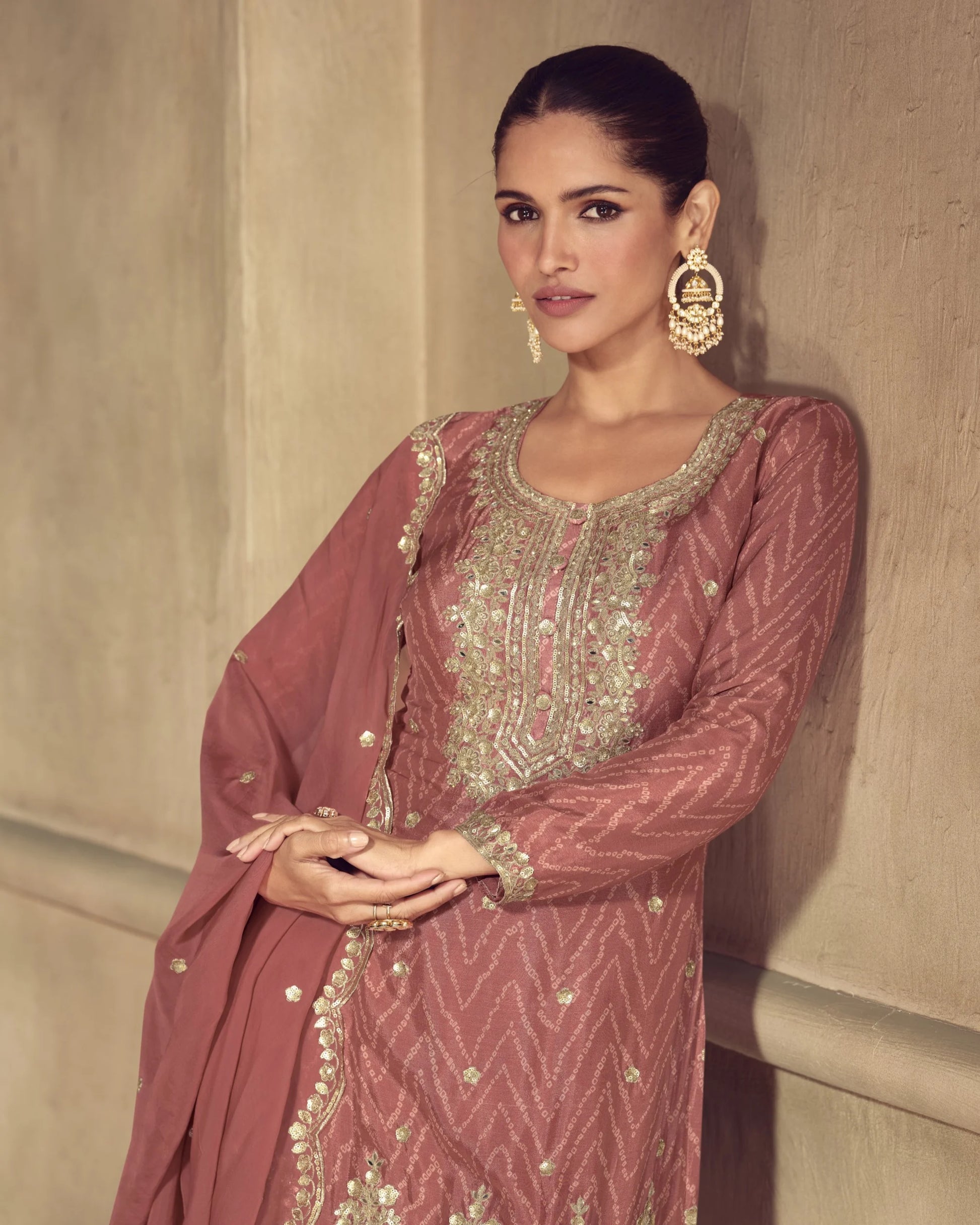 Woman wearing a traditional pink and gold embroidered outfit against a beige wall.