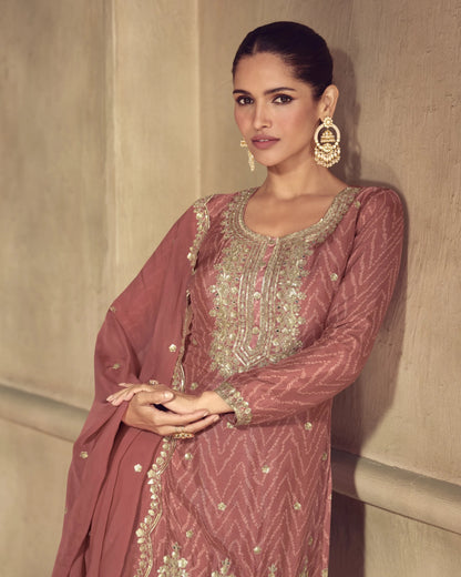 Woman wearing a traditional pink and gold embroidered outfit against a beige wall.