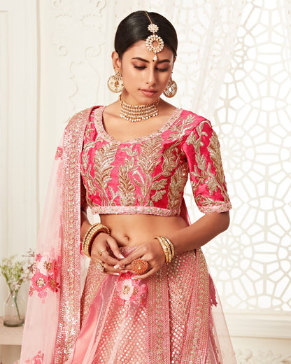 Woman wearing a pink and gold embroidered traditional outfit with jewelry against a decorative white wall.