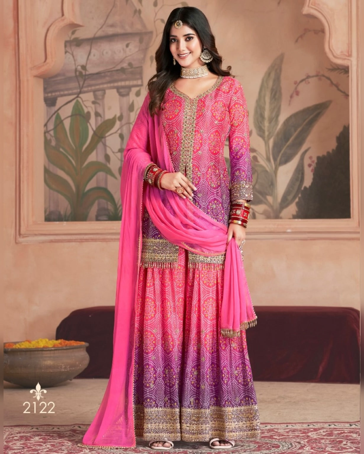 Woman in a pink and purple traditional outfit with a matching dupatta against a decorative wall.