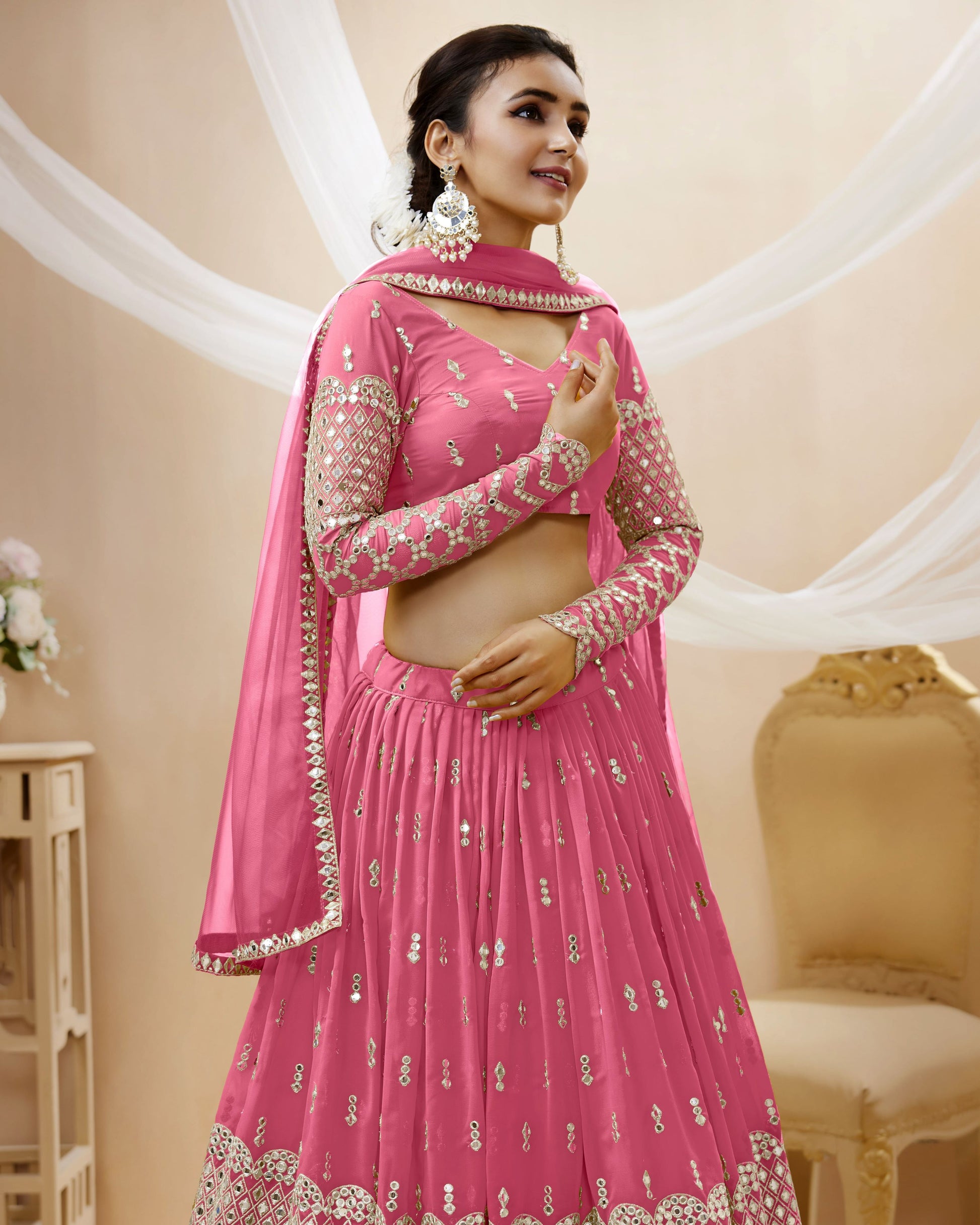 Woman wearing a pink traditional outfit with intricate designs in a decorated room.