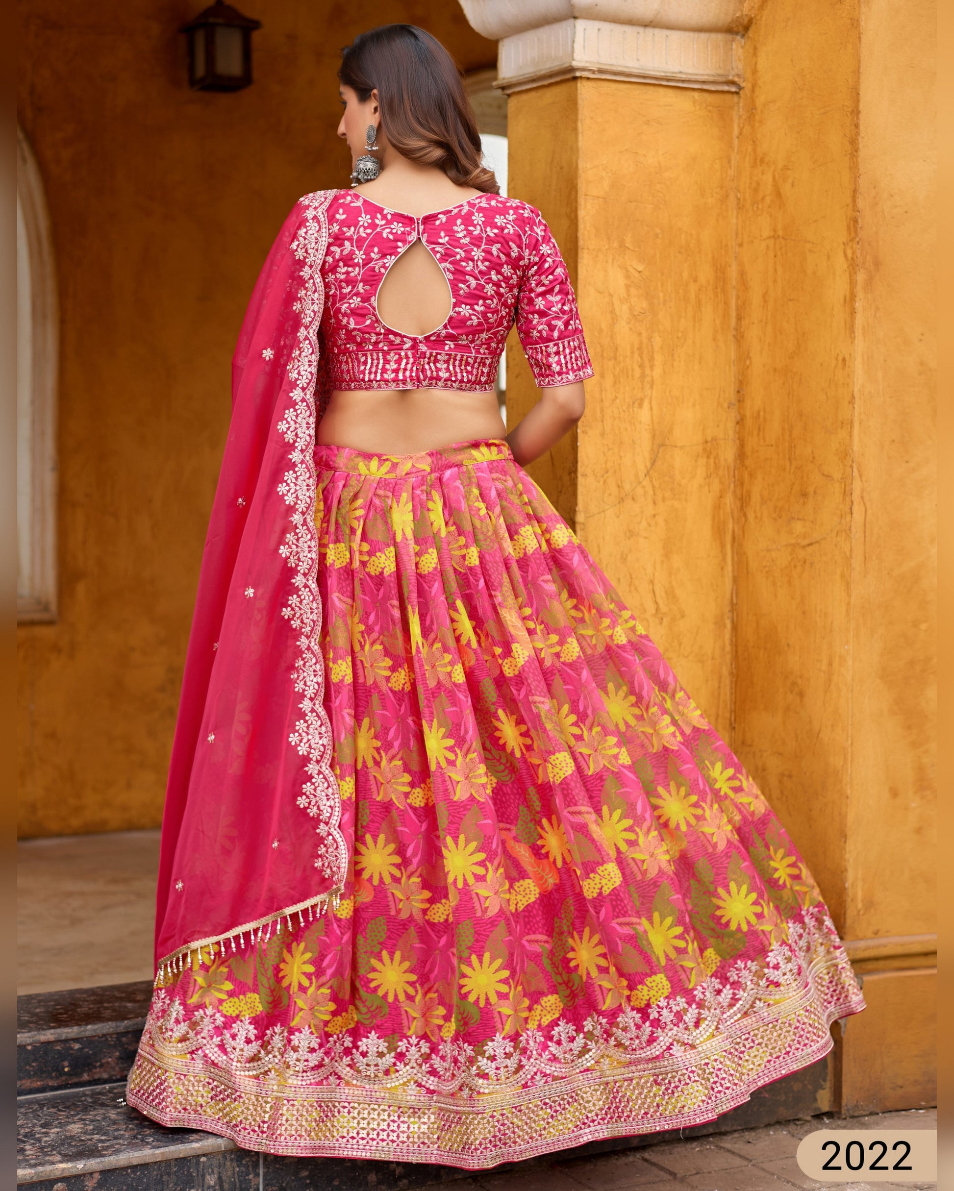 Woman wearing a pink and yellow traditional outfit with a floral pattern, standing against a warm-toned wall.