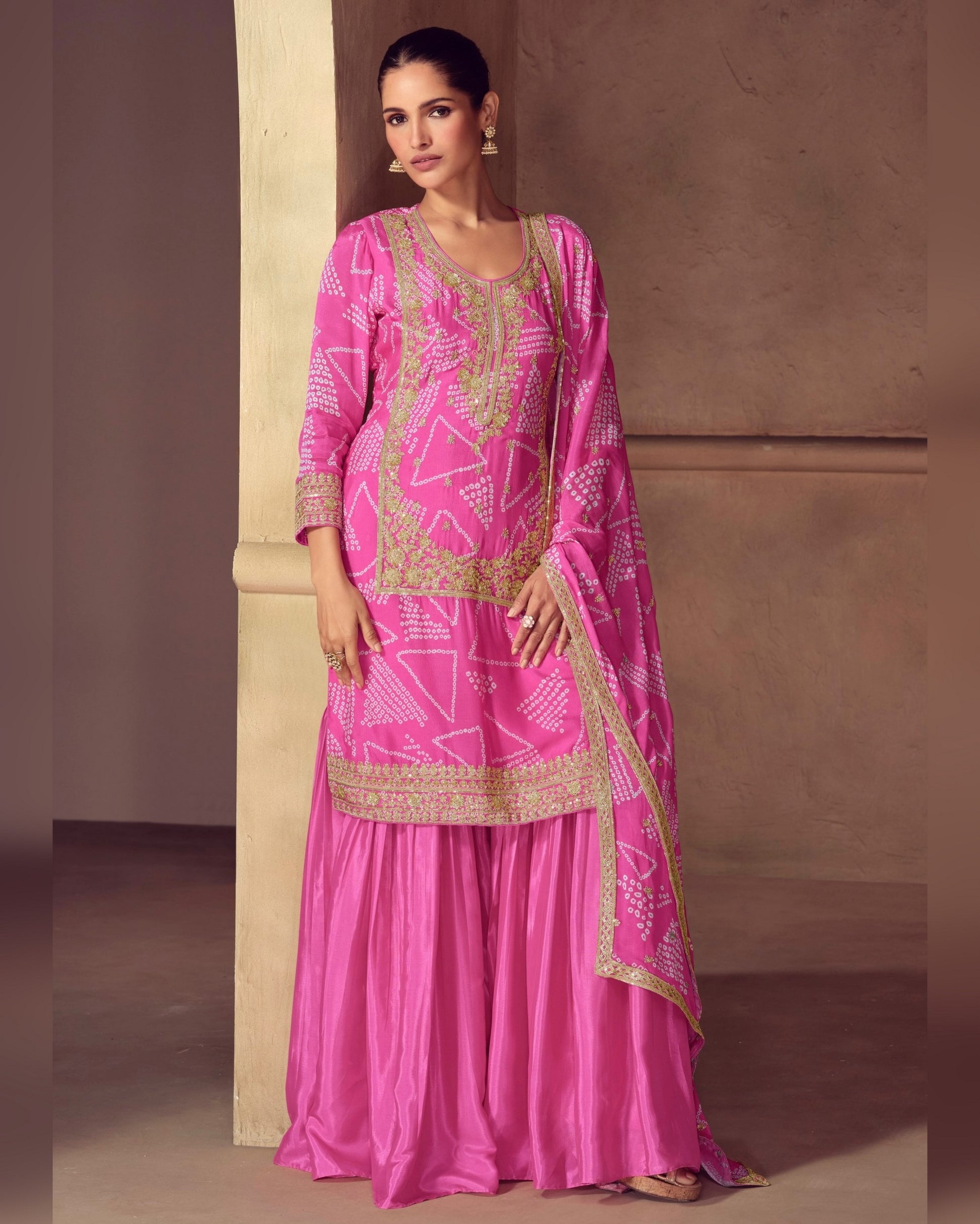 Woman wearing a pink traditional outfit with gold embroidery against a beige wall.