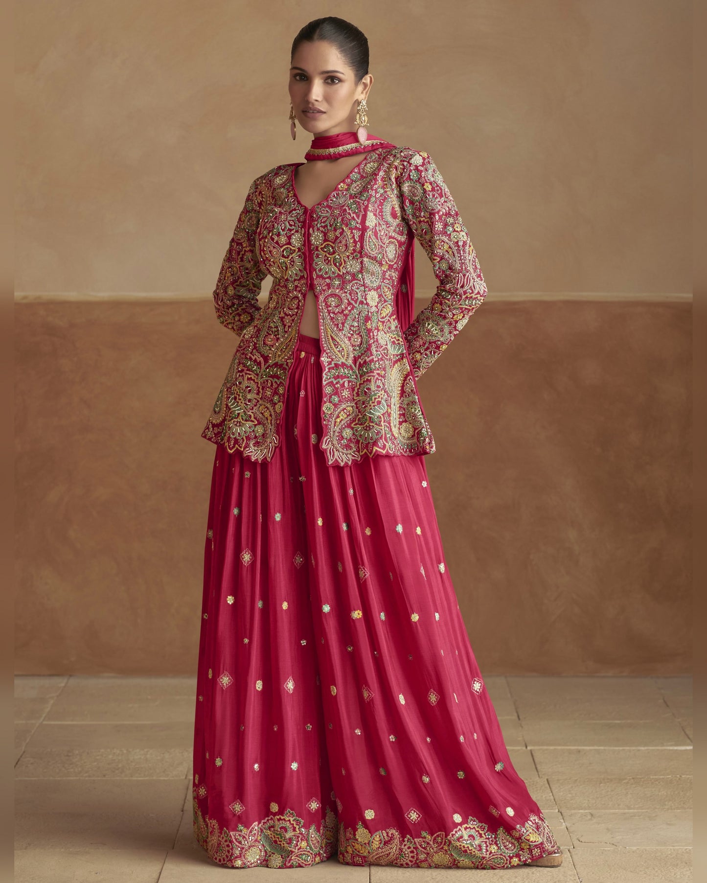 Woman wearing a traditional outfit with a pink lehenga and embroidered jacket against a beige wall.