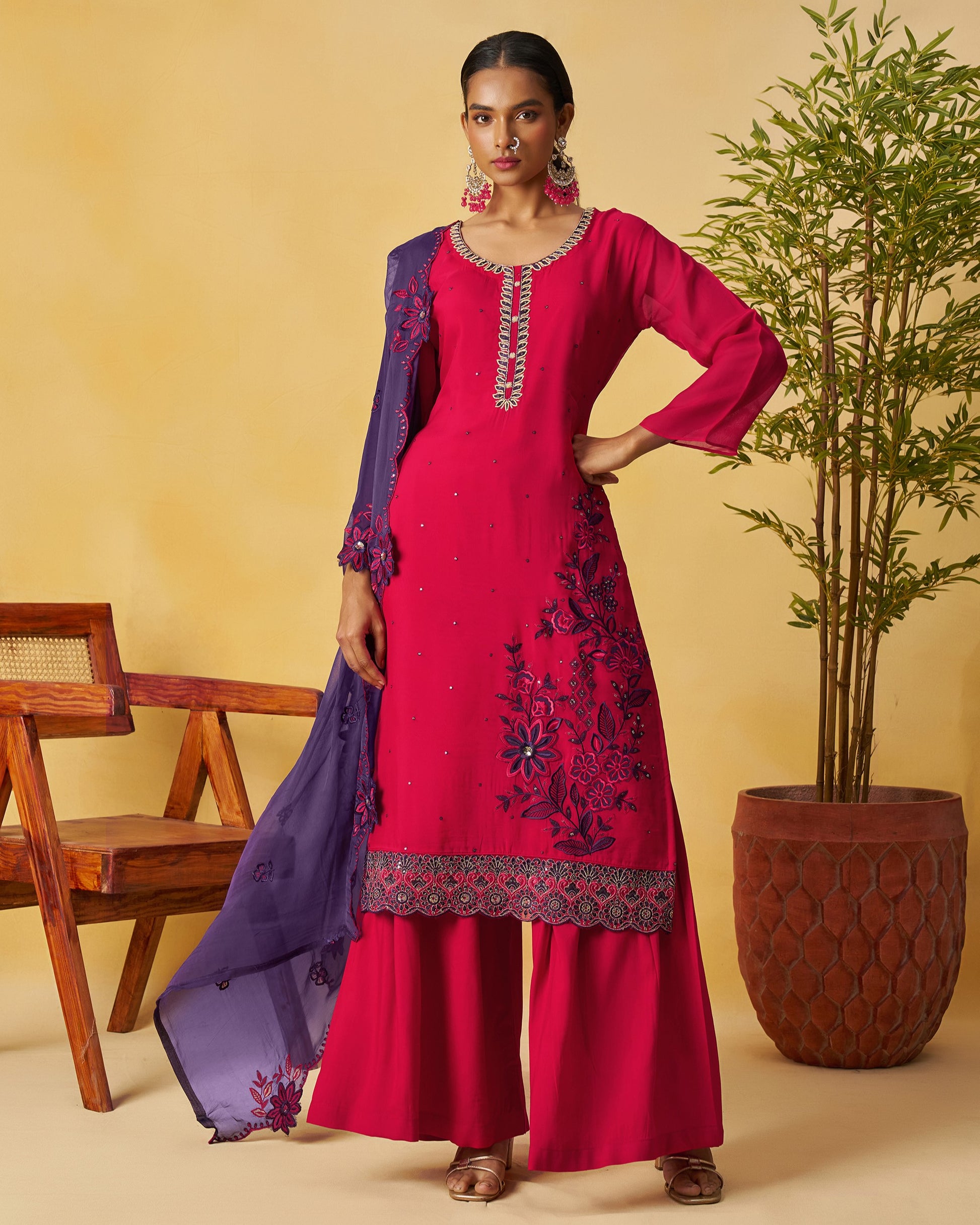 Woman in a red embroidered outfit with a purple dupatta against a beige background