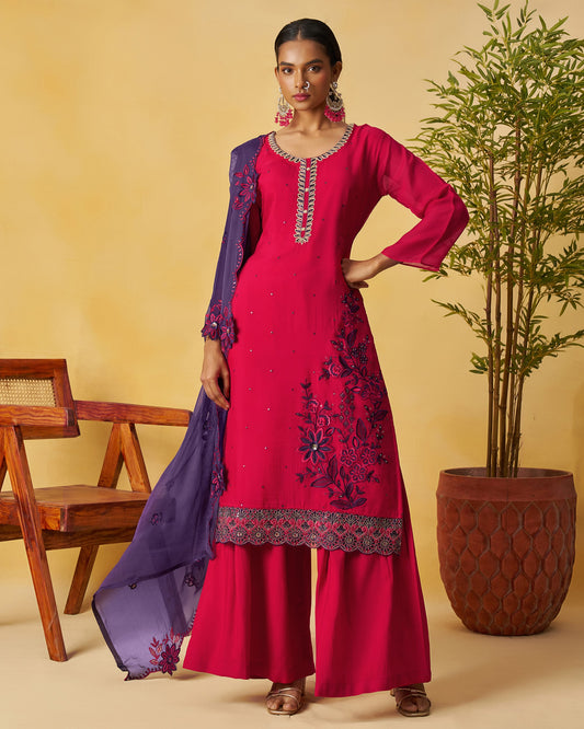 Woman in a red embroidered outfit with a purple dupatta against a beige background