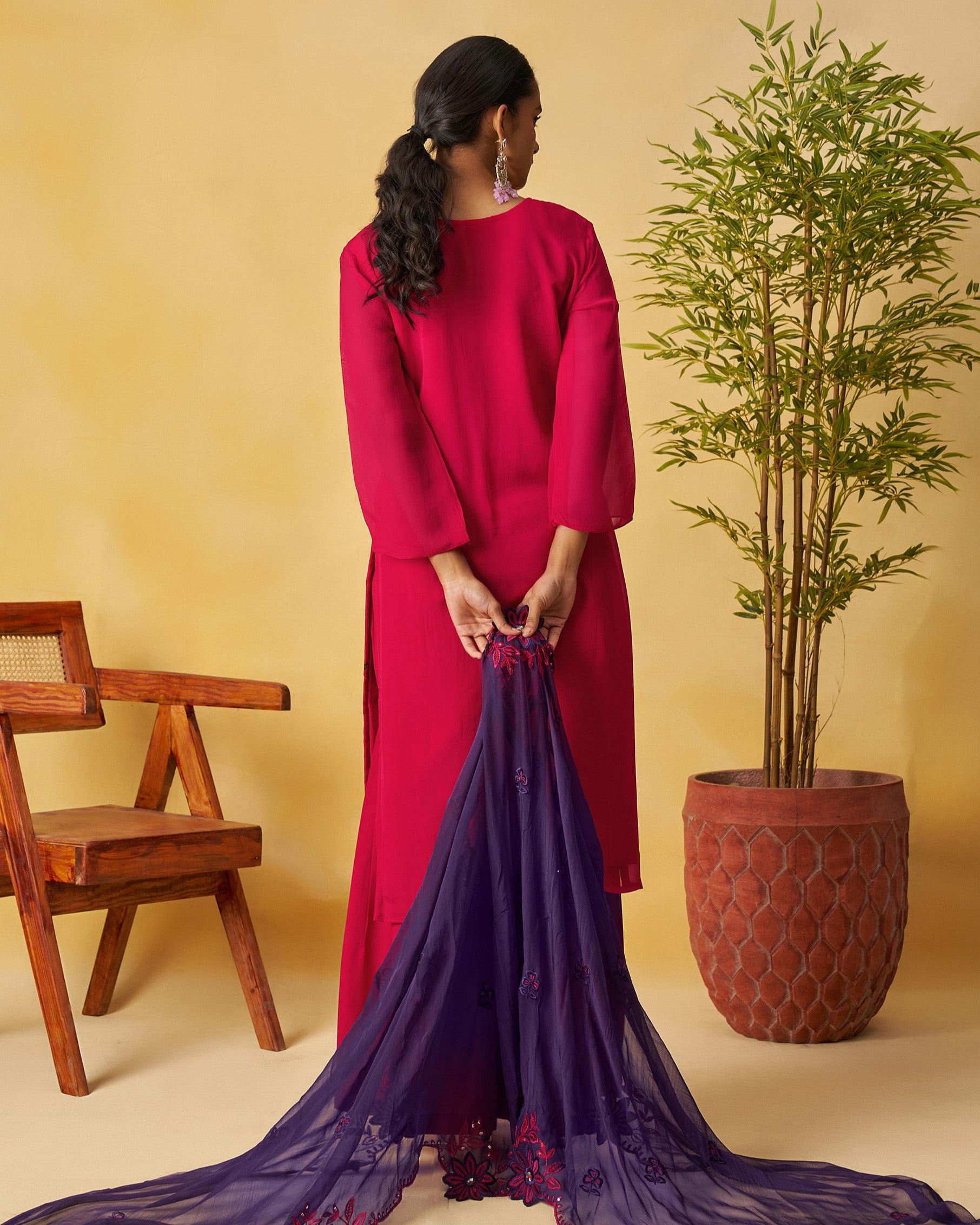 Woman in a red dress holding a long, dark blue fabric against a beige wall with a wooden chair and potted plant.