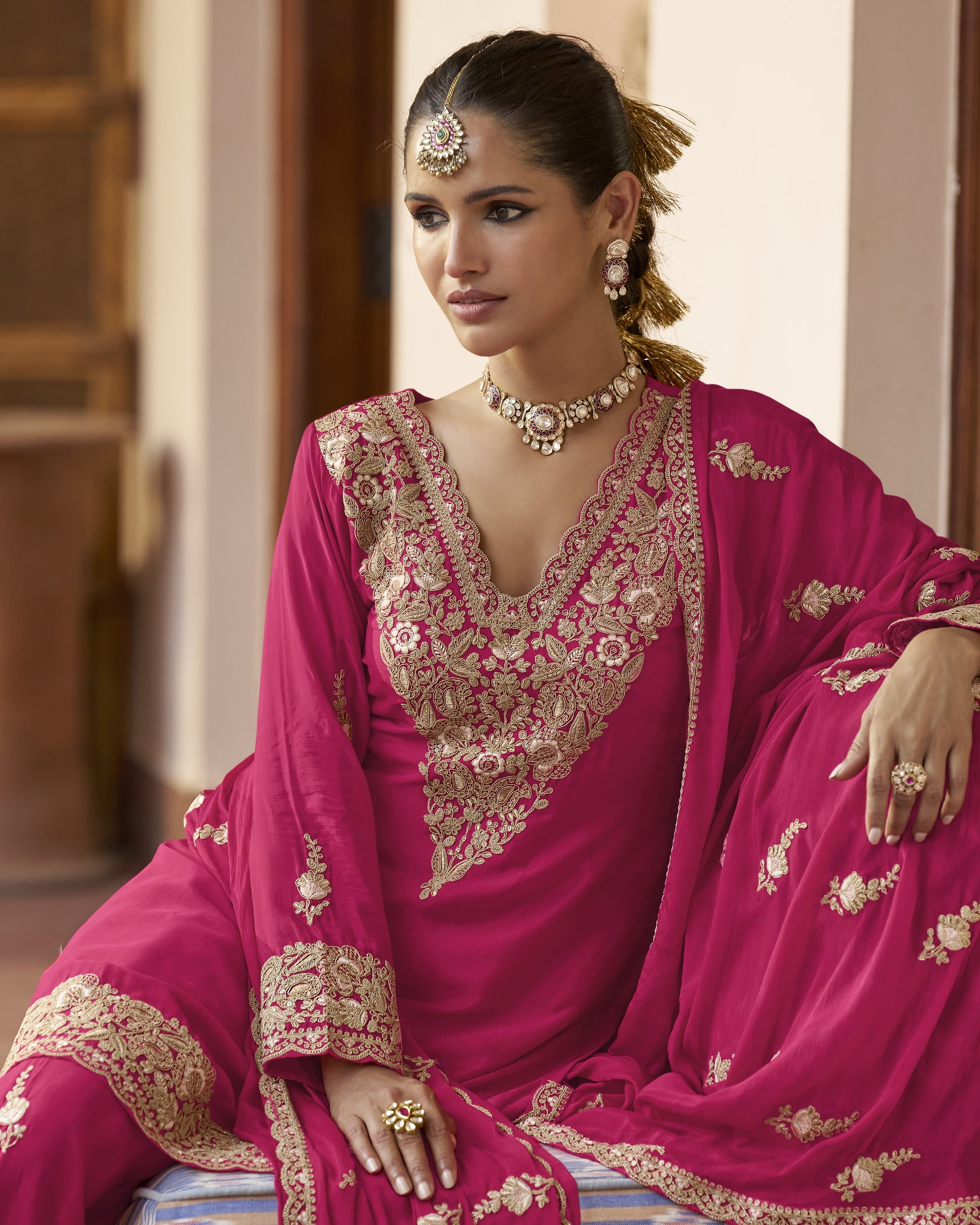 Woman in a pink embroidered traditional outfit with gold jewelry indoors.