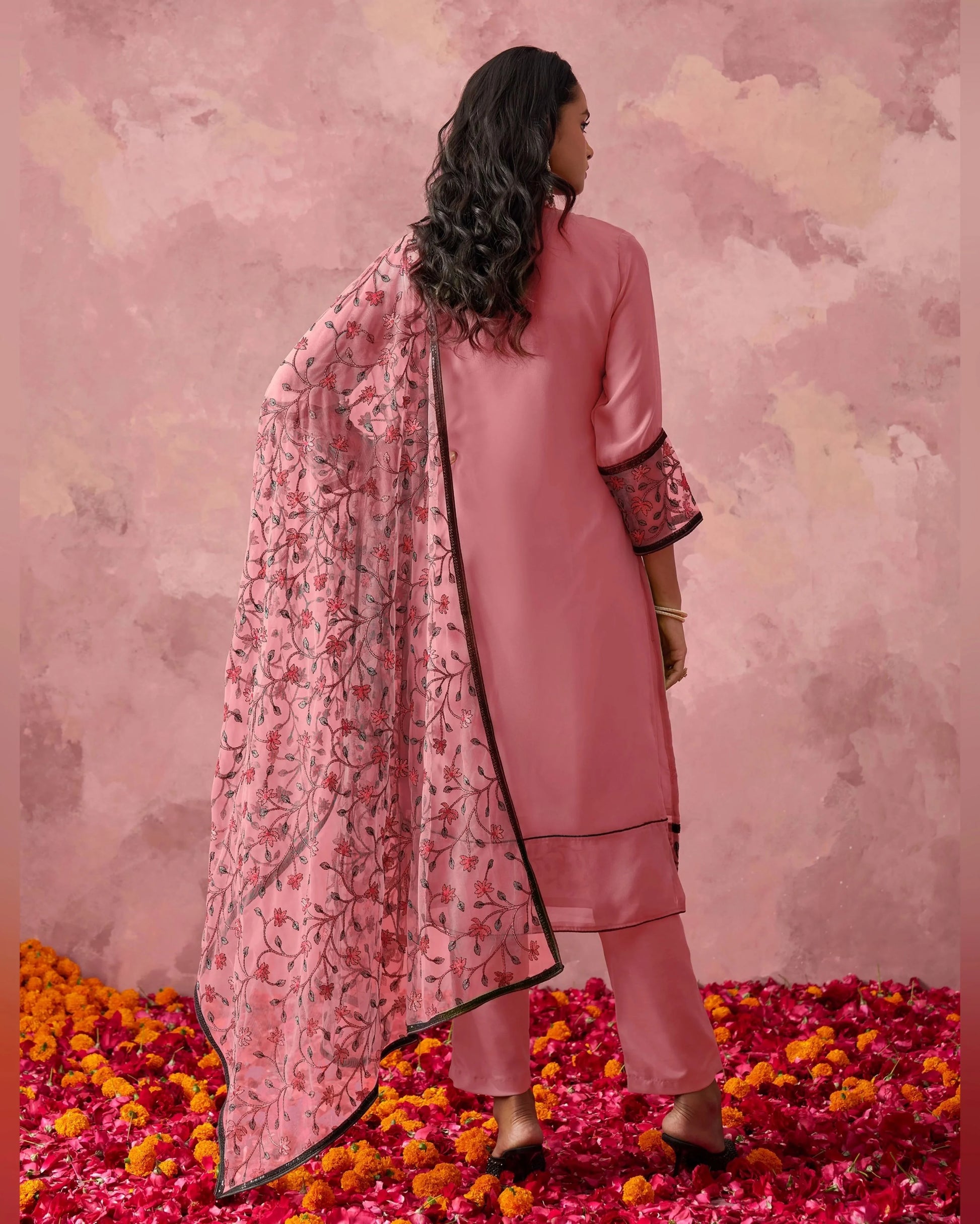 Woman in a pink traditional outfit with a floral dupatta standing on rose petals against a pink background