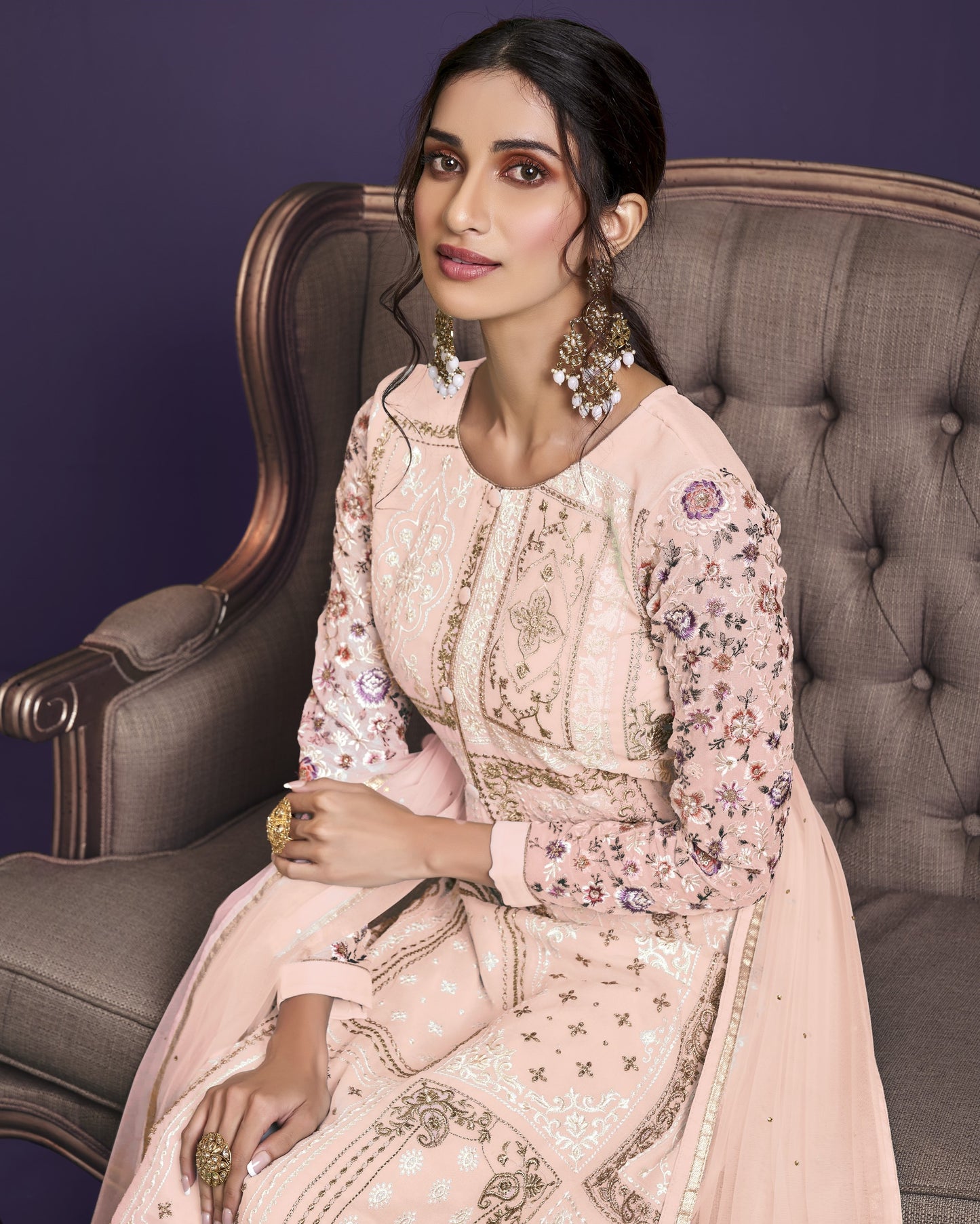 Woman in a pink embroidered outfit sitting on a gray chair against a purple background