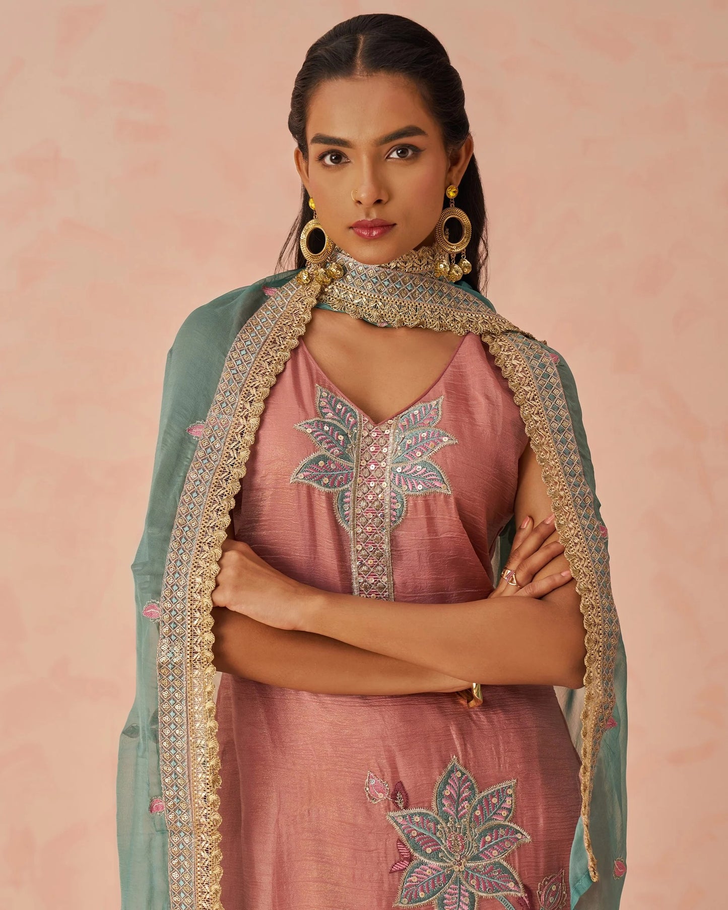 Woman wearing a traditional outfit with pink top and pants and green dupatta against a pink background