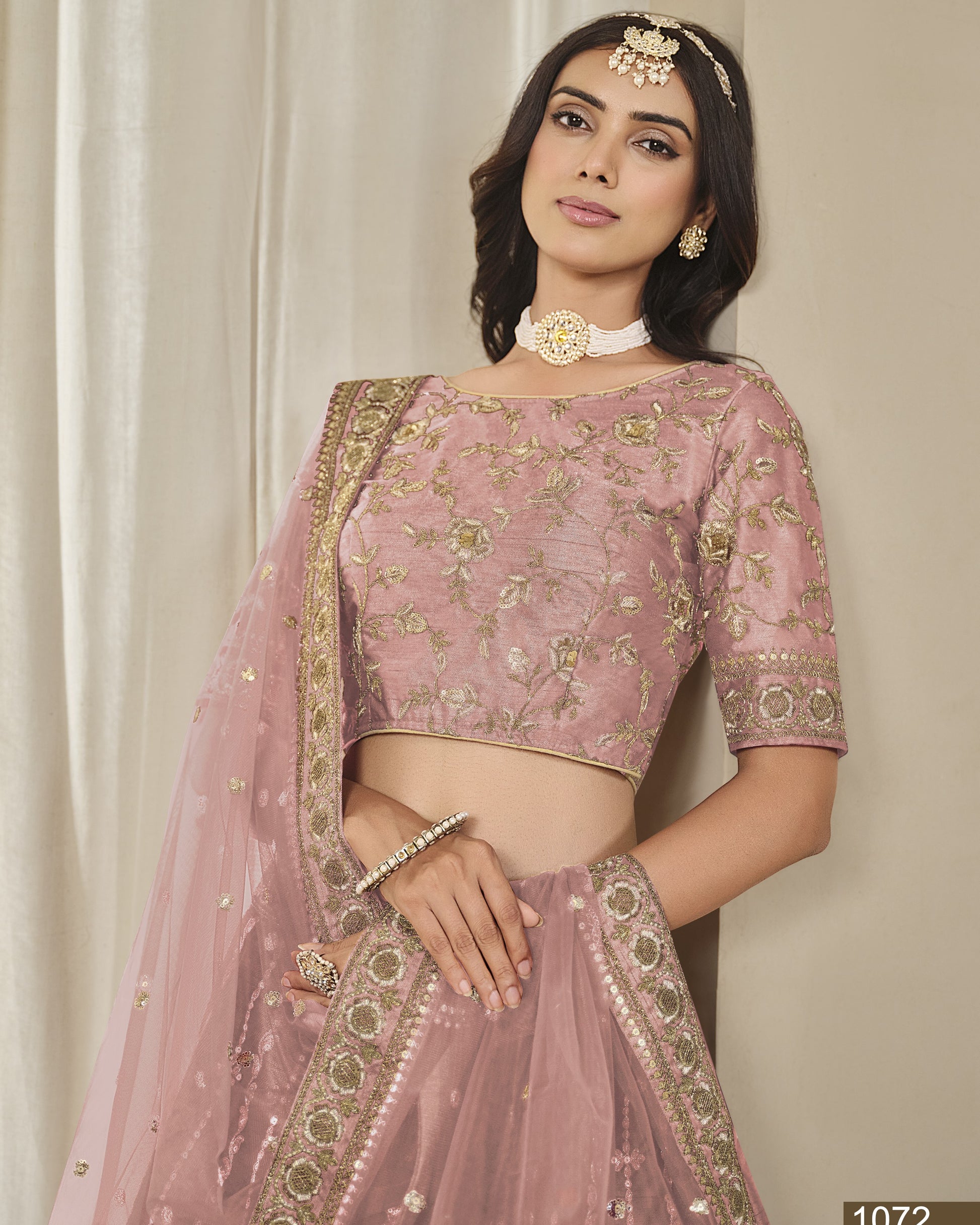 Woman wearing a pink embroidered saree with gold details against a white curtain background