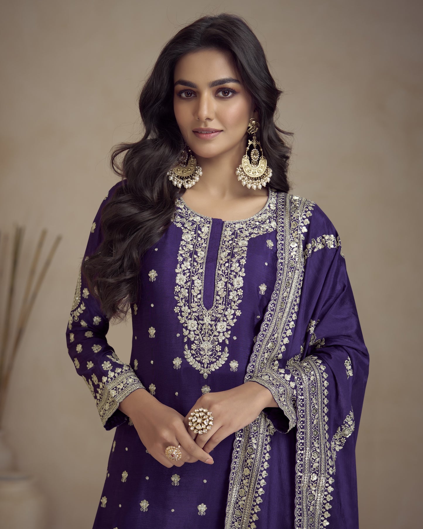 Woman wearing a purple embroidered traditional outfit with gold jewelry against a neutral background