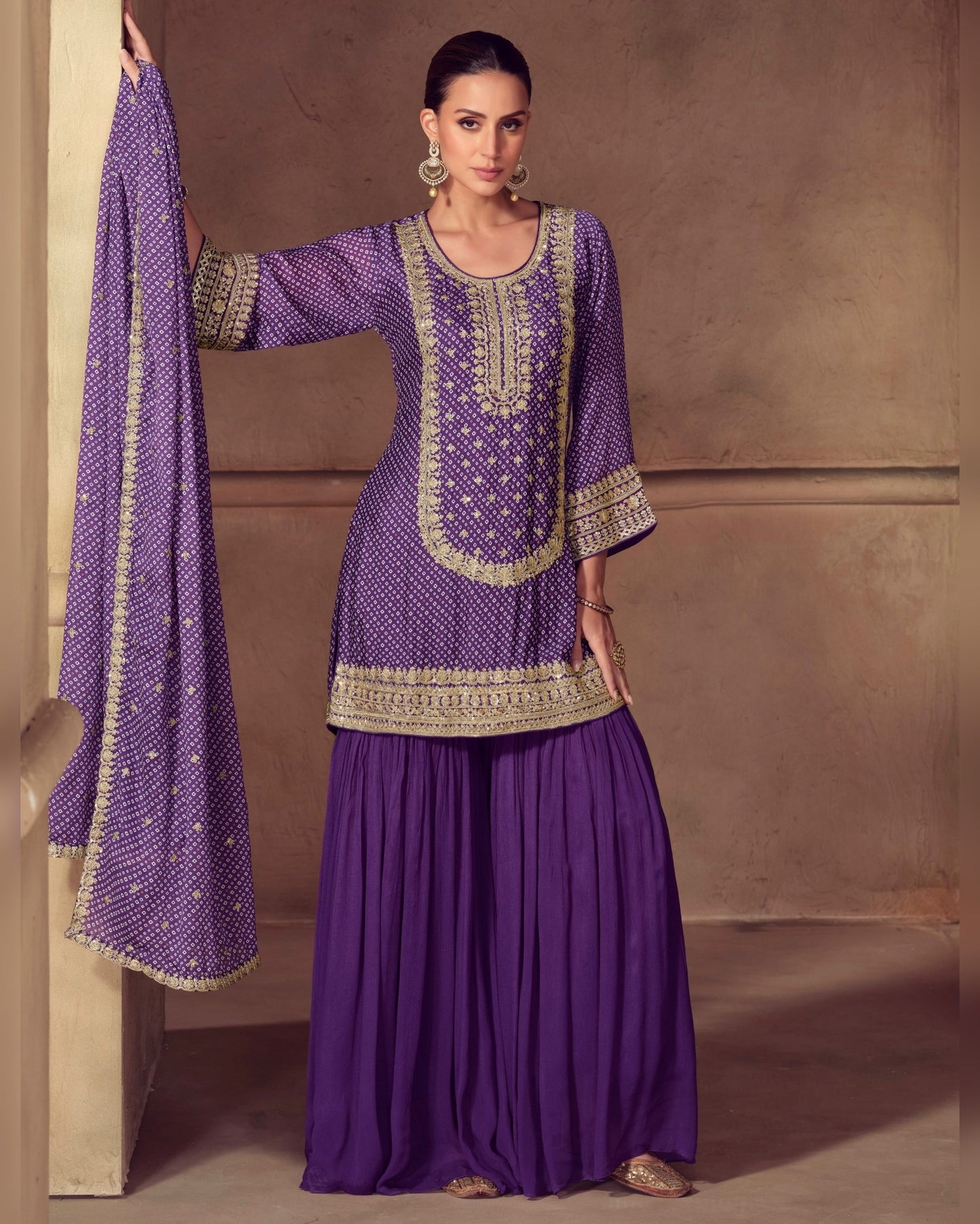 Woman wearing a purple traditional outfit with gold embroidery against a beige wall.