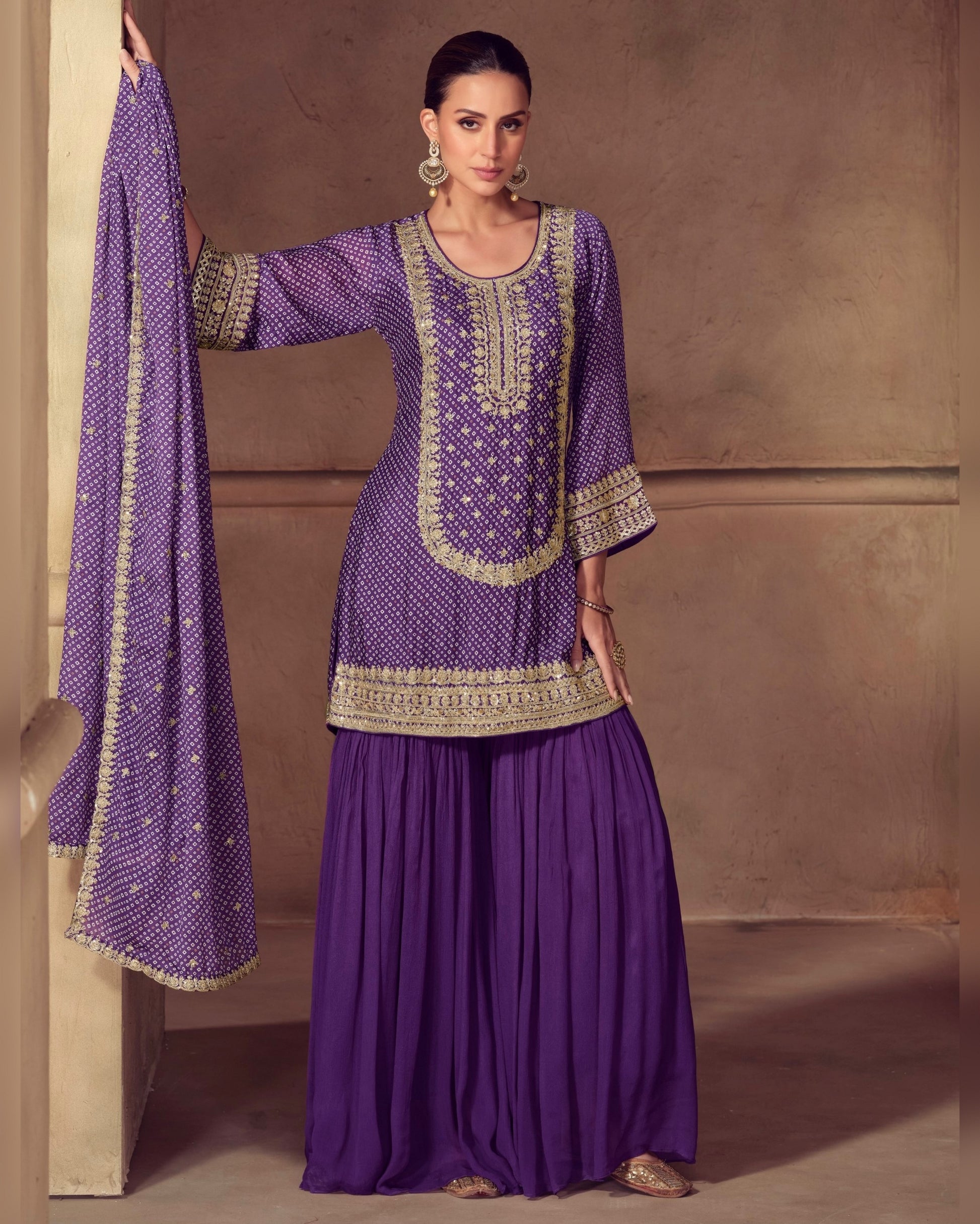 Woman wearing a purple traditional outfit with gold embroidery against a beige wall.