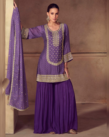 Woman wearing a purple traditional outfit with gold embroidery against a beige wall.