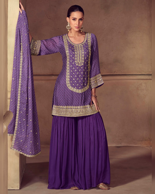 Woman wearing a purple traditional outfit with gold embroidery against a beige wall.