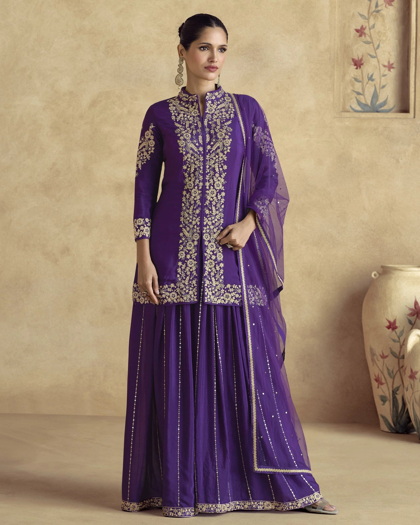 Woman wearing a purple traditional outfit with gold embroidery against a beige wall.