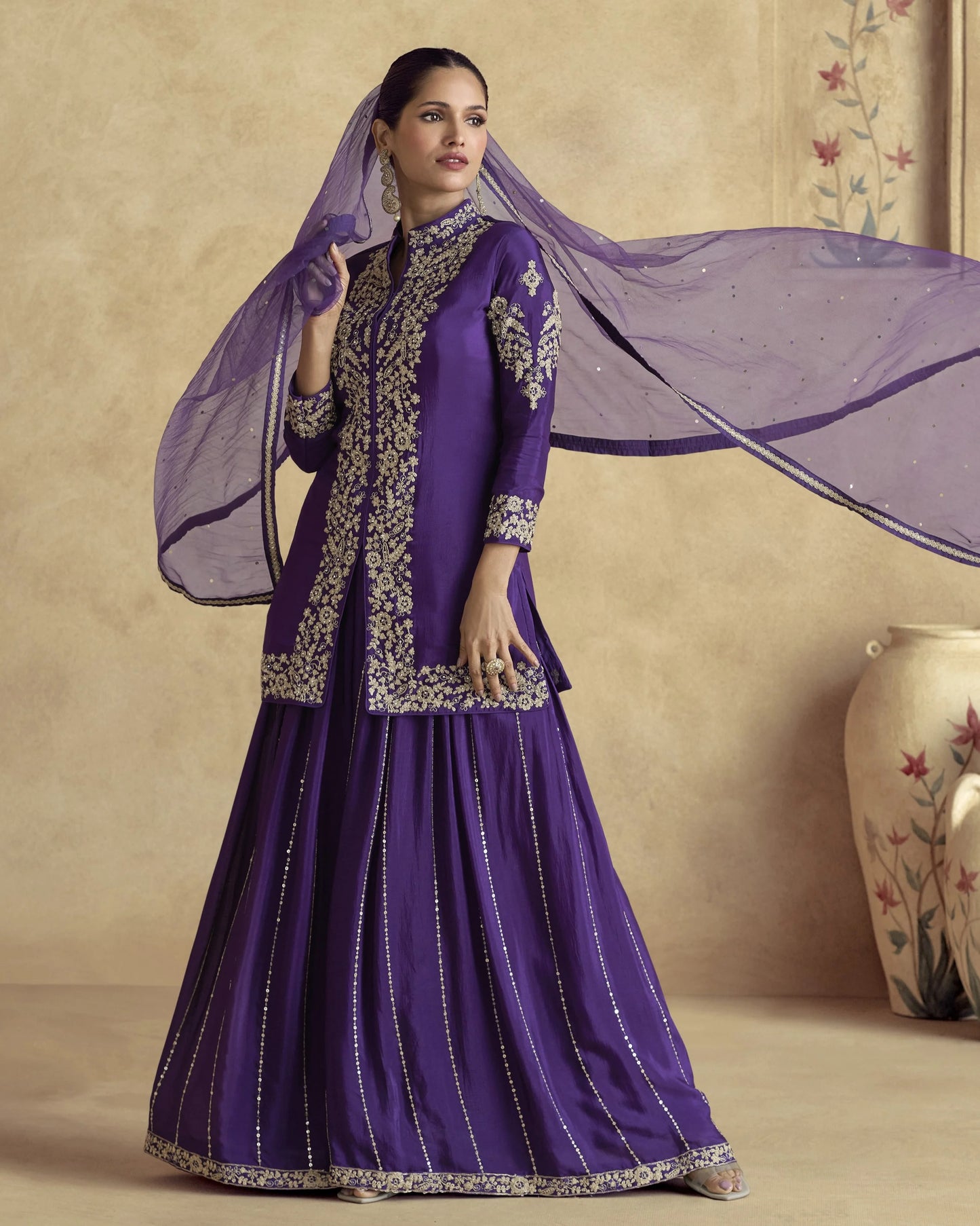 Woman in a purple traditional outfit with a sheer dupatta against a beige wall.