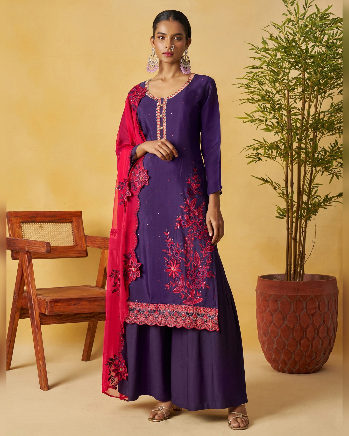 Woman in a purple traditional outfit with red embroidery standing next to a wooden chair and potted plant.
