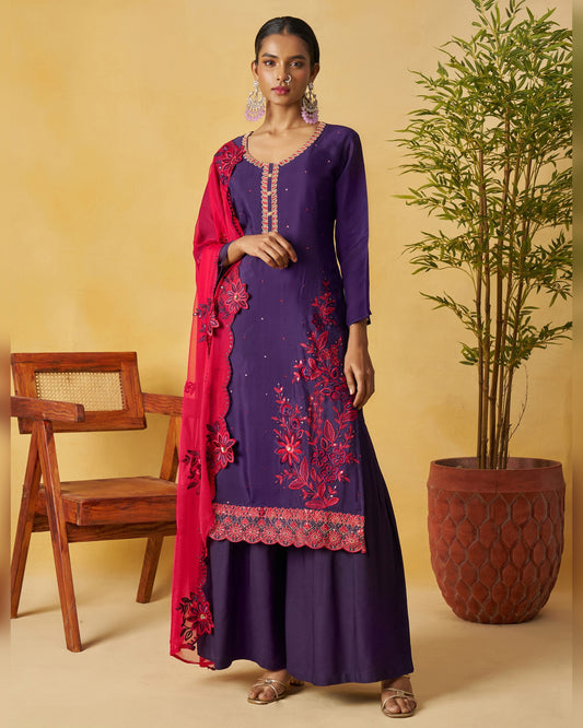 Woman in a purple traditional outfit with red embroidery standing next to a wooden chair and potted plant.