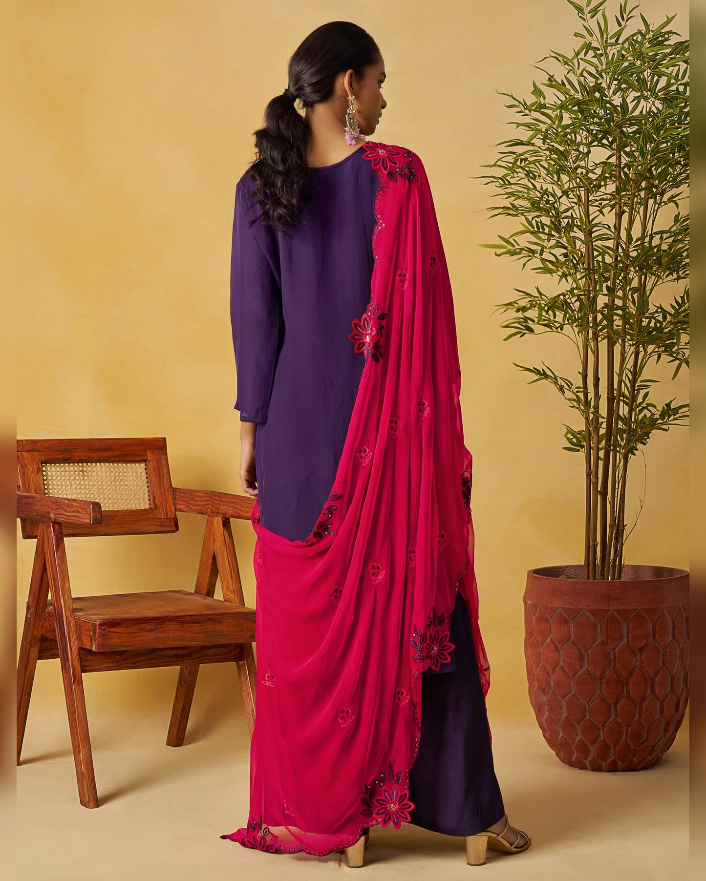 Woman in a purple top and pink saree standing next to a wooden chair and potted plant against a beige wall.