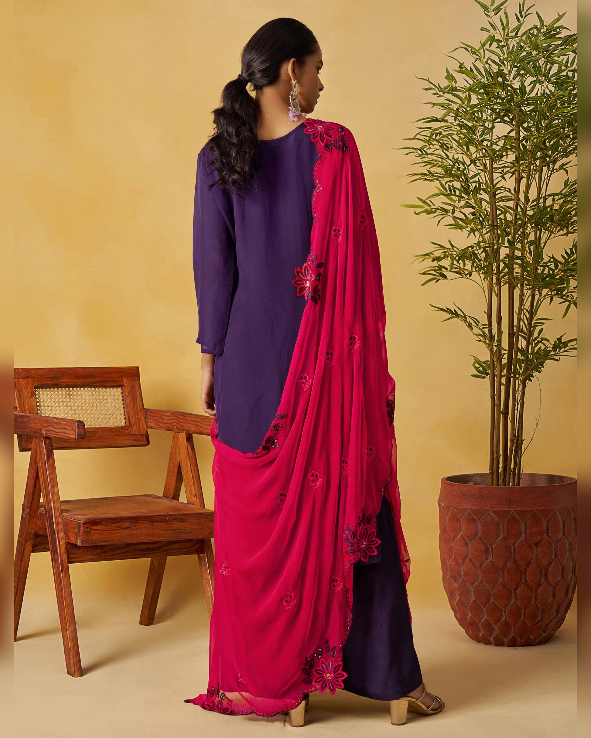 Woman in a purple top and pink saree standing next to a wooden chair and potted plant against a beige wall.
