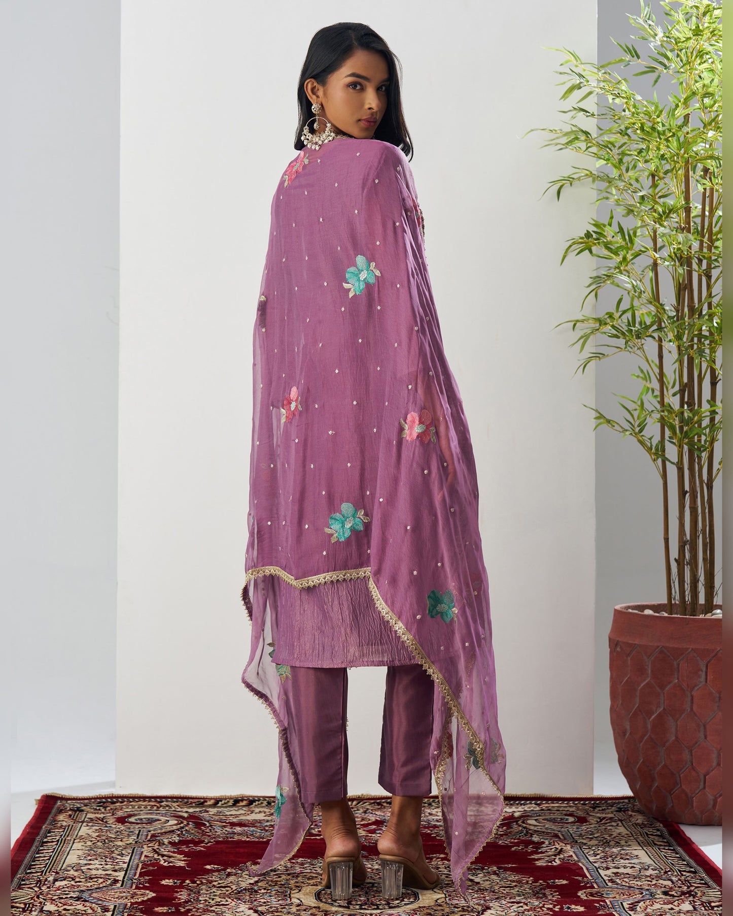 Woman wearing a purple embroidered outfit standing in a room with a plant and patterned rug.