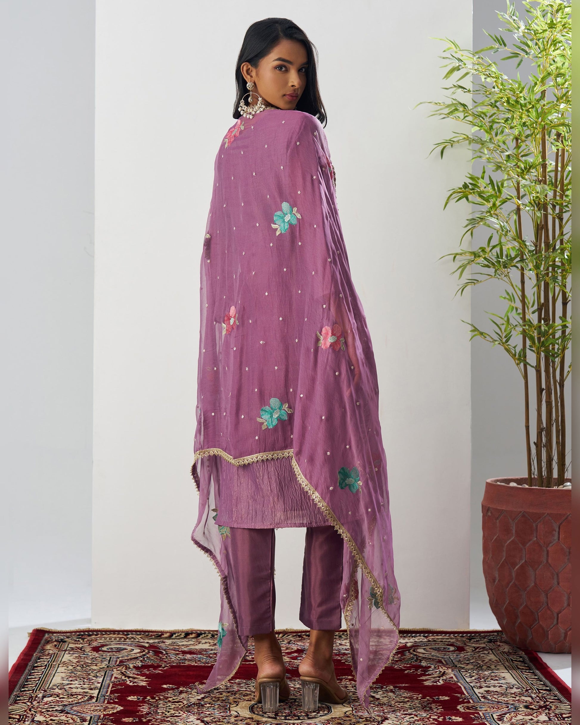 Woman wearing a purple embroidered outfit standing in a room with a plant and patterned rug.