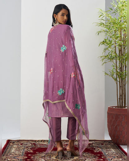 Woman wearing a purple embroidered outfit standing in a room with a plant and patterned rug.