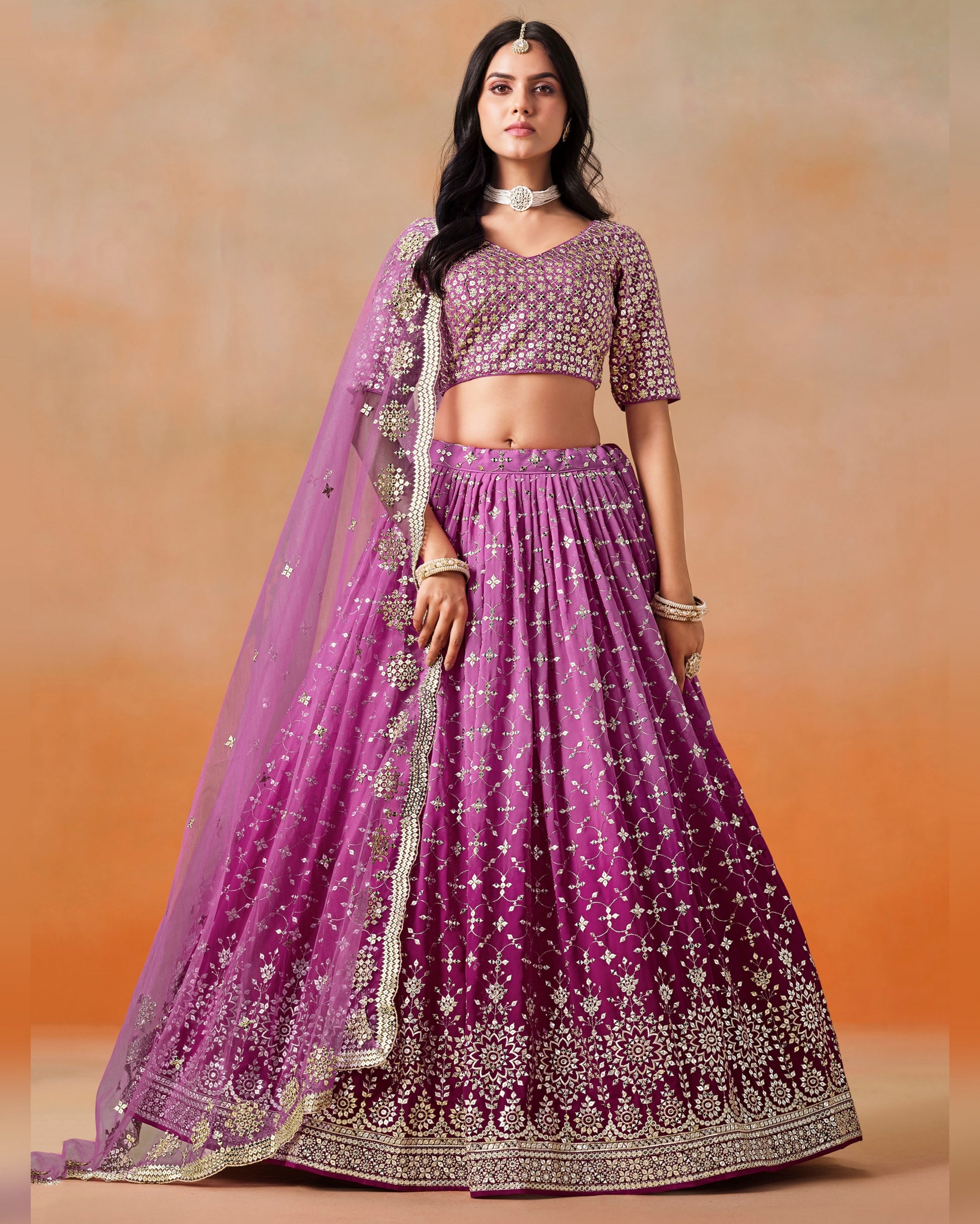 Woman wearing a purple embroidered traditional outfit with a matching dupatta on an orange background