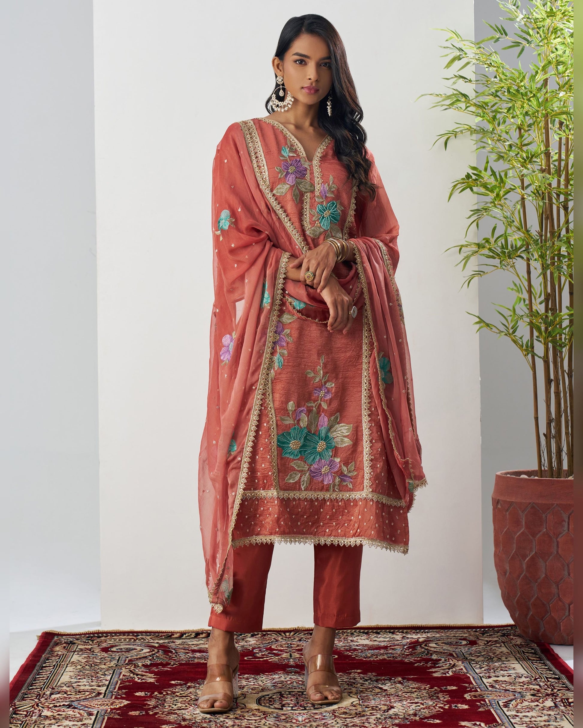 Woman wearing a coral pink embroidered traditional outfit with floral patterns on a plain background